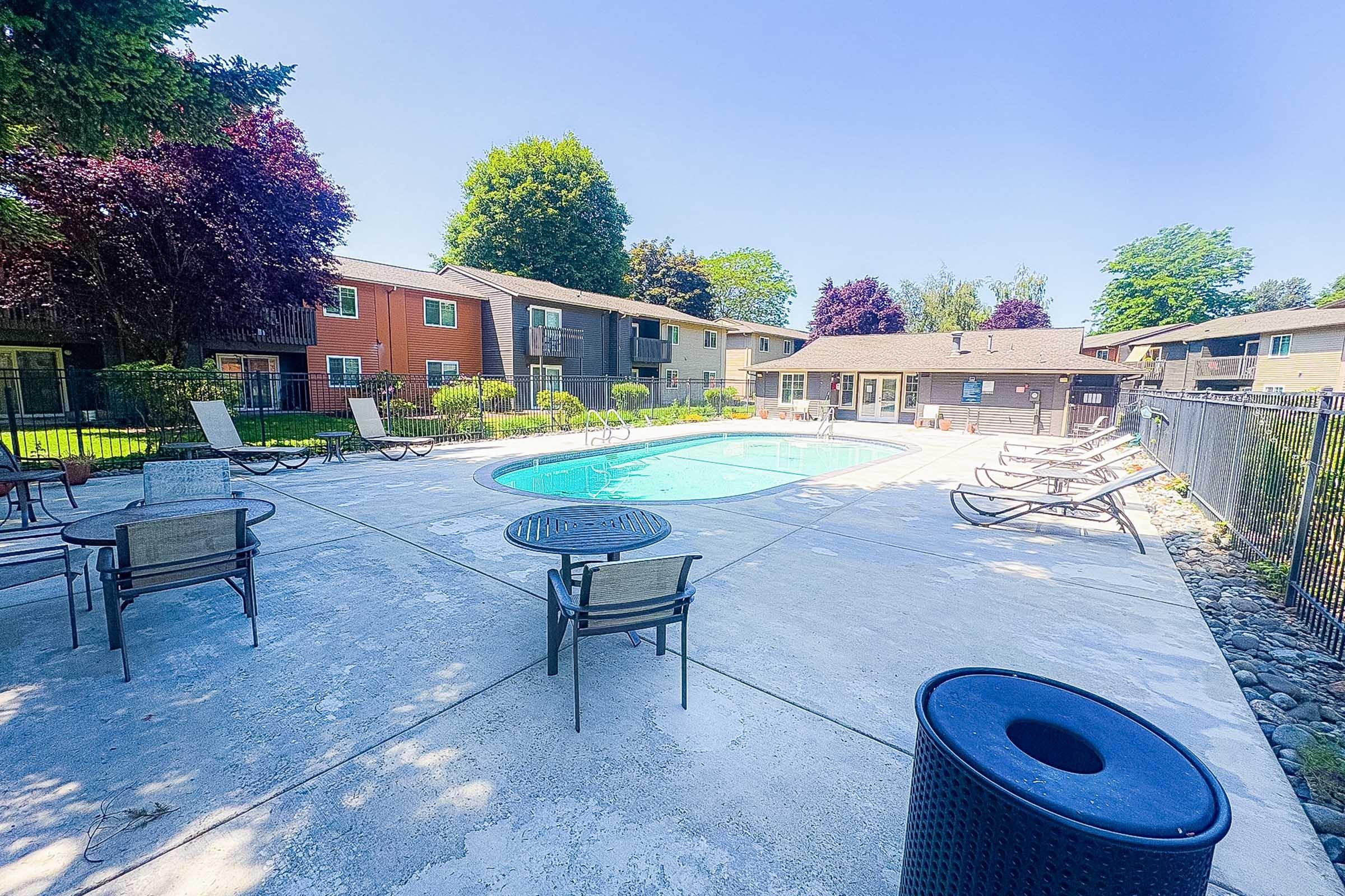 A sparkling blue swimming pool surrounded by lounge chairs and a concrete deck, situated in a residential area with green trees and colorful buildings in the background. Clear skies enhance the inviting atmosphere of the pool area.