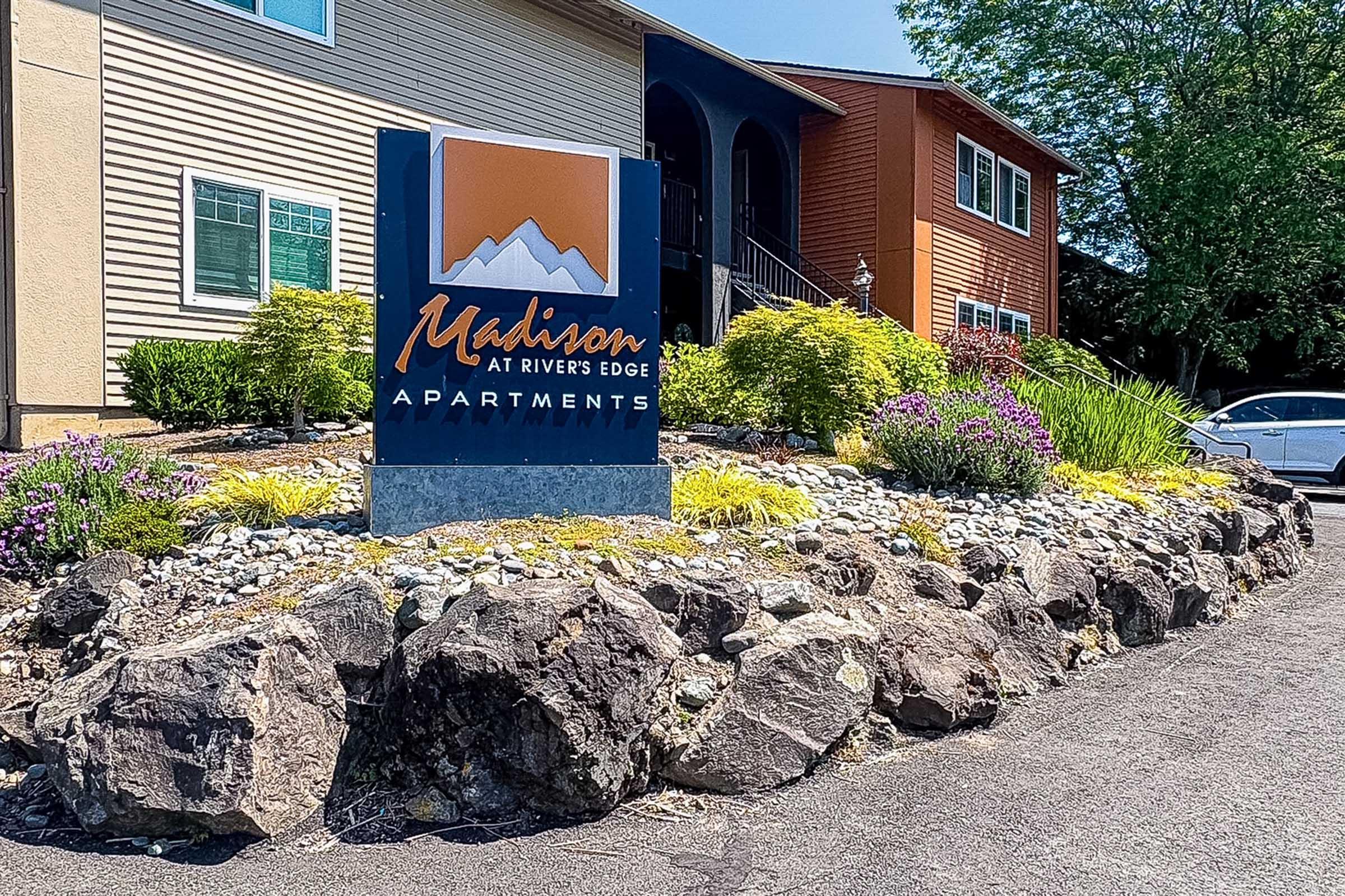 Signboard for Madison at River's Edge Apartments, featuring an orange and blue logo with mountains. Surrounded by landscaped greenery, including flowers and shrubs, with rocks and a pathway leading to the entrance of a multi-story apartment building. Bright, sunny day.