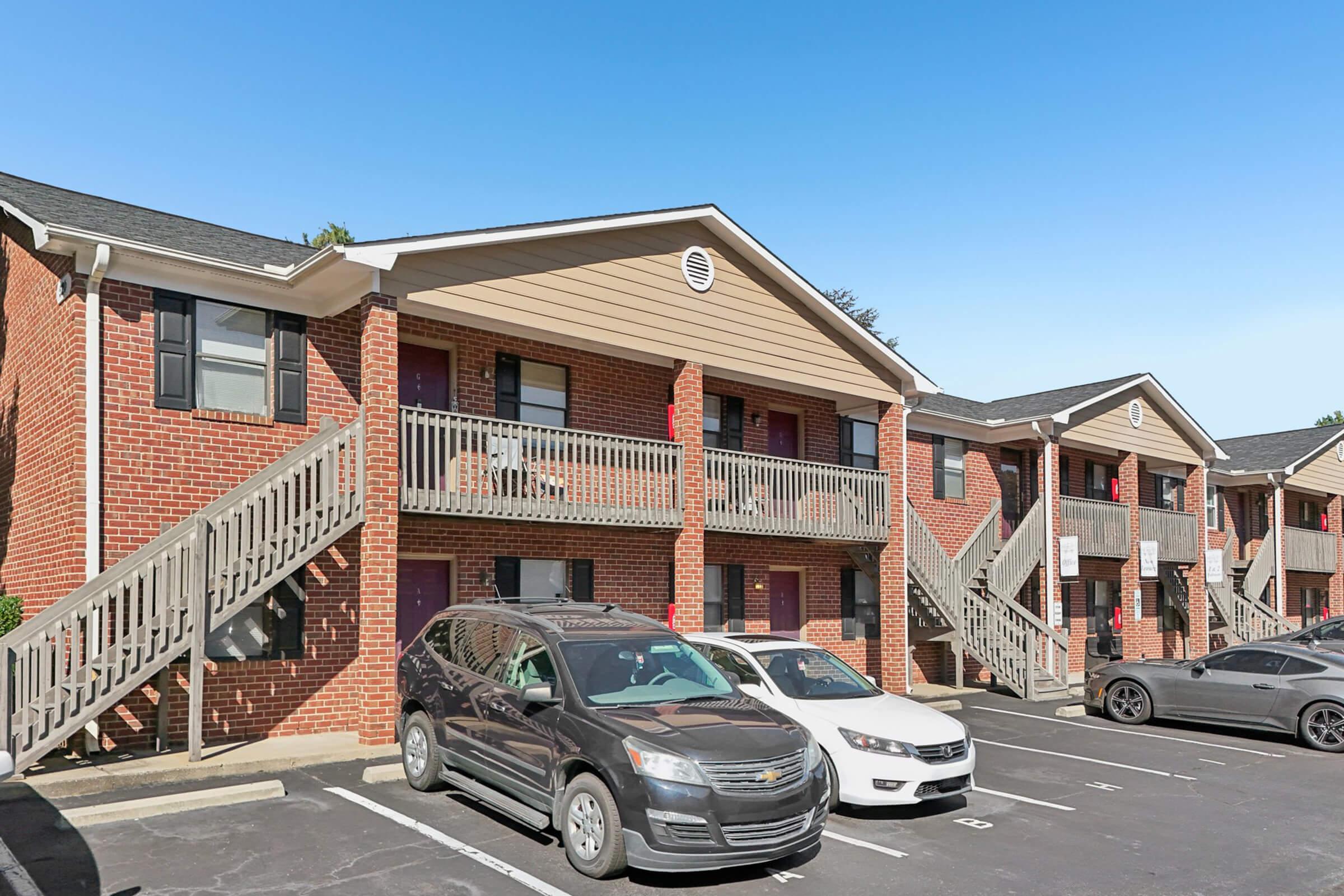 Row of brick apartment buildings with wooden staircases and balconies, set against a clear blue sky. Several parked cars are visible in front, showcasing a mix of colors and models. The setting appears to be a residential area with ample parking space.