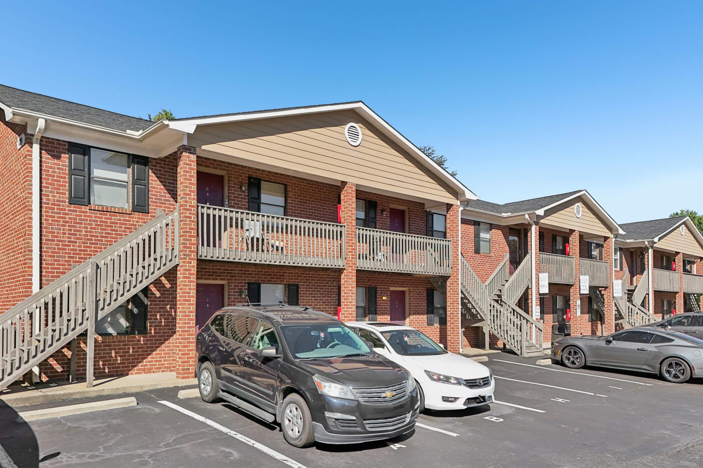 A two-story brick apartment building with multiple units, featuring stairs leading to balconies on the second floor. The parking lot is visible in front, with several parked cars, including a gray SUV and a white sedan. The sky is clear and blue, creating a bright atmosphere.