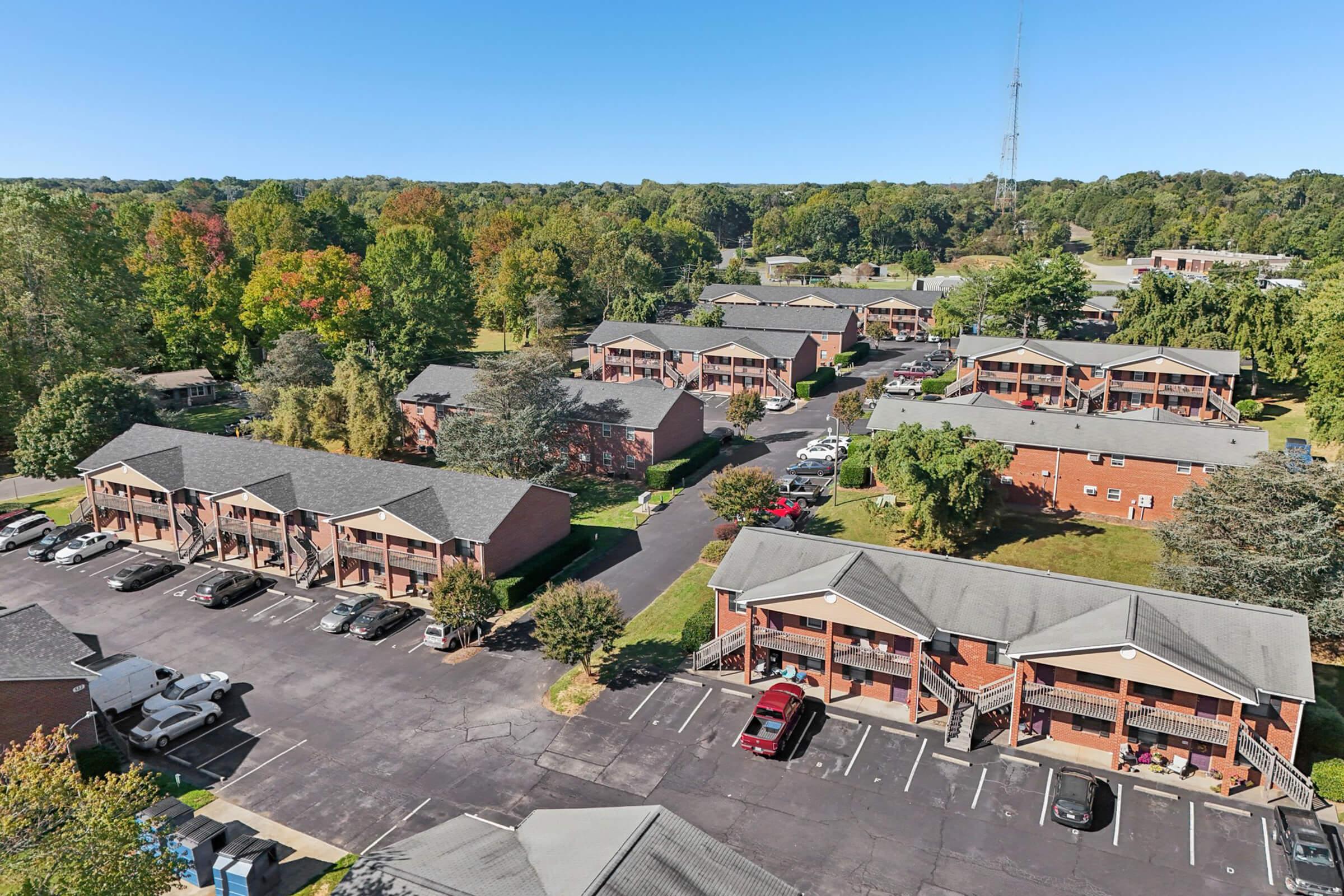 Aerial view of a residential complex featuring several brick apartment buildings arranged around a parking area. The surrounding landscape includes trees with varying autumn colors, and a tall radio tower in the distance under a clear blue sky.