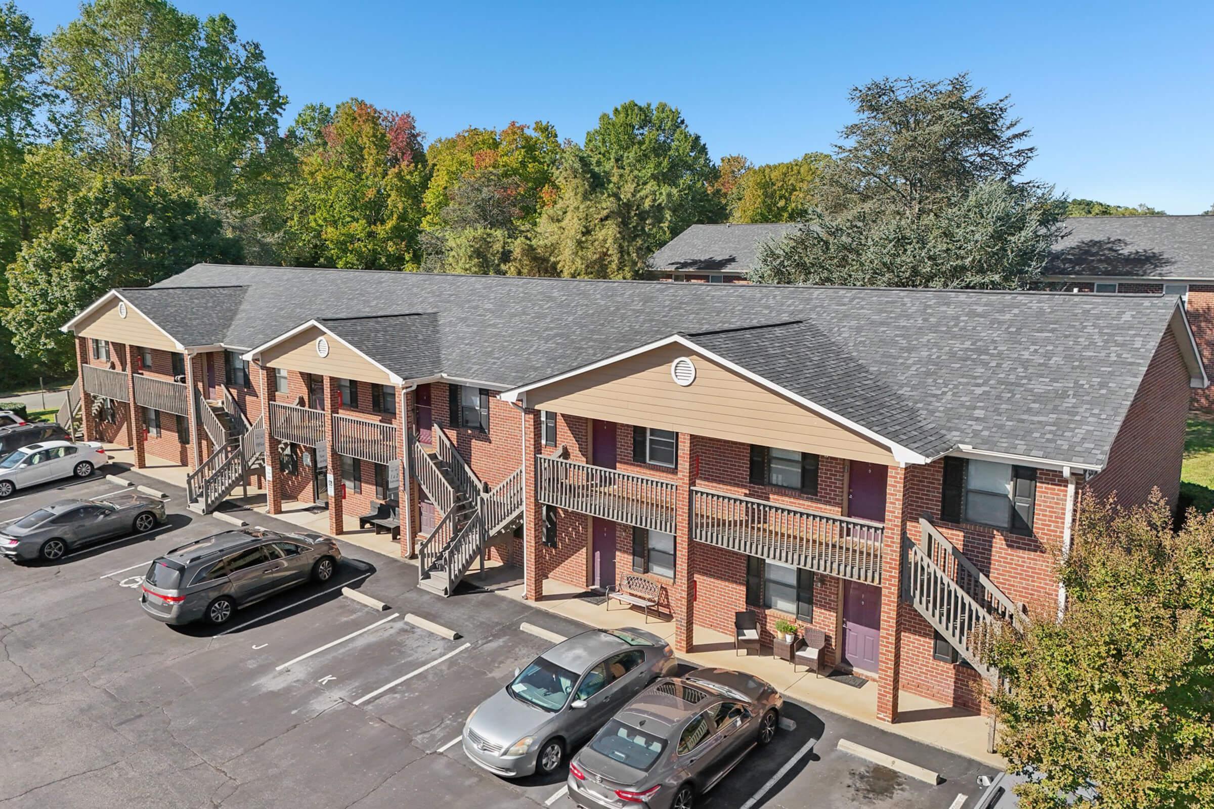 Aerial view of a brick apartment building with multiple units featuring balconies. Surrounding trees show autumn colors, and several parked cars are visible in the foreground. The sky is clear and blue, providing a bright backdrop to the scene.