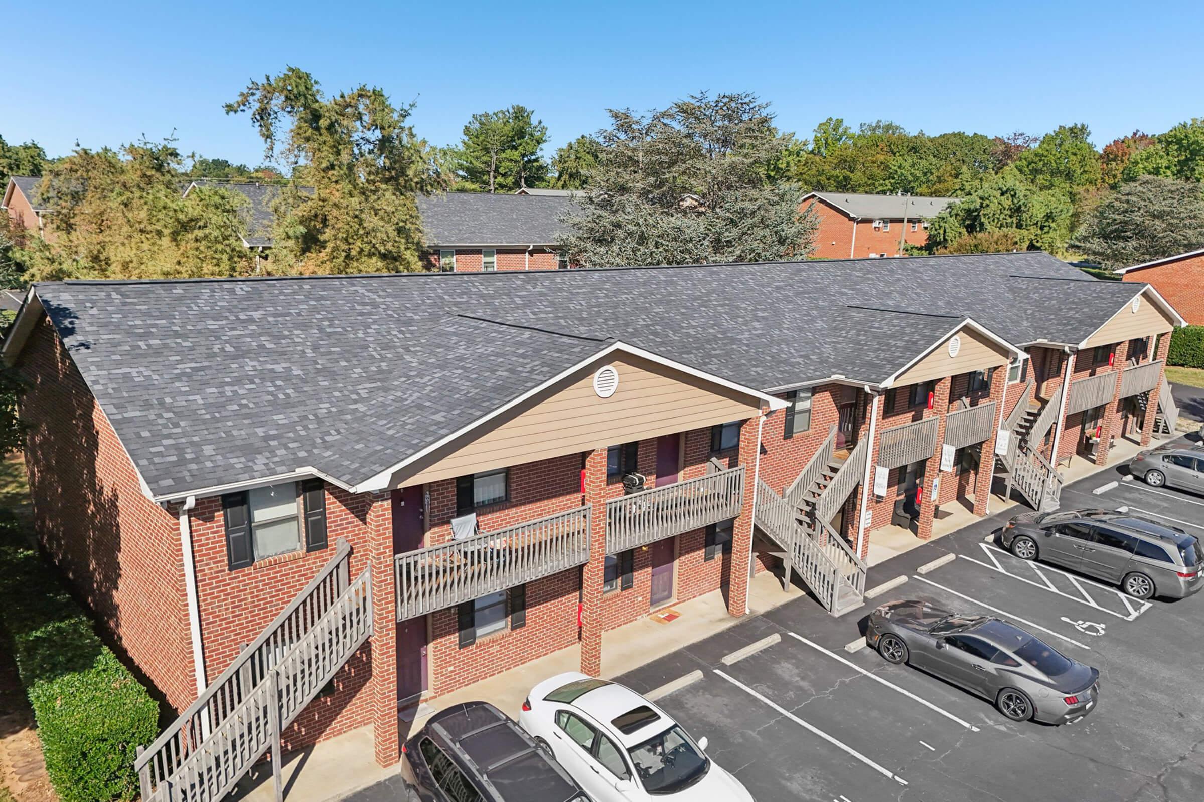 View of a two-story brick apartment complex featuring multiple units with balconies. The roofs are dark gray shingles, and there are several parked cars in front of the buildings. The surrounding area has trees and lush greenery, under a clear blue sky.