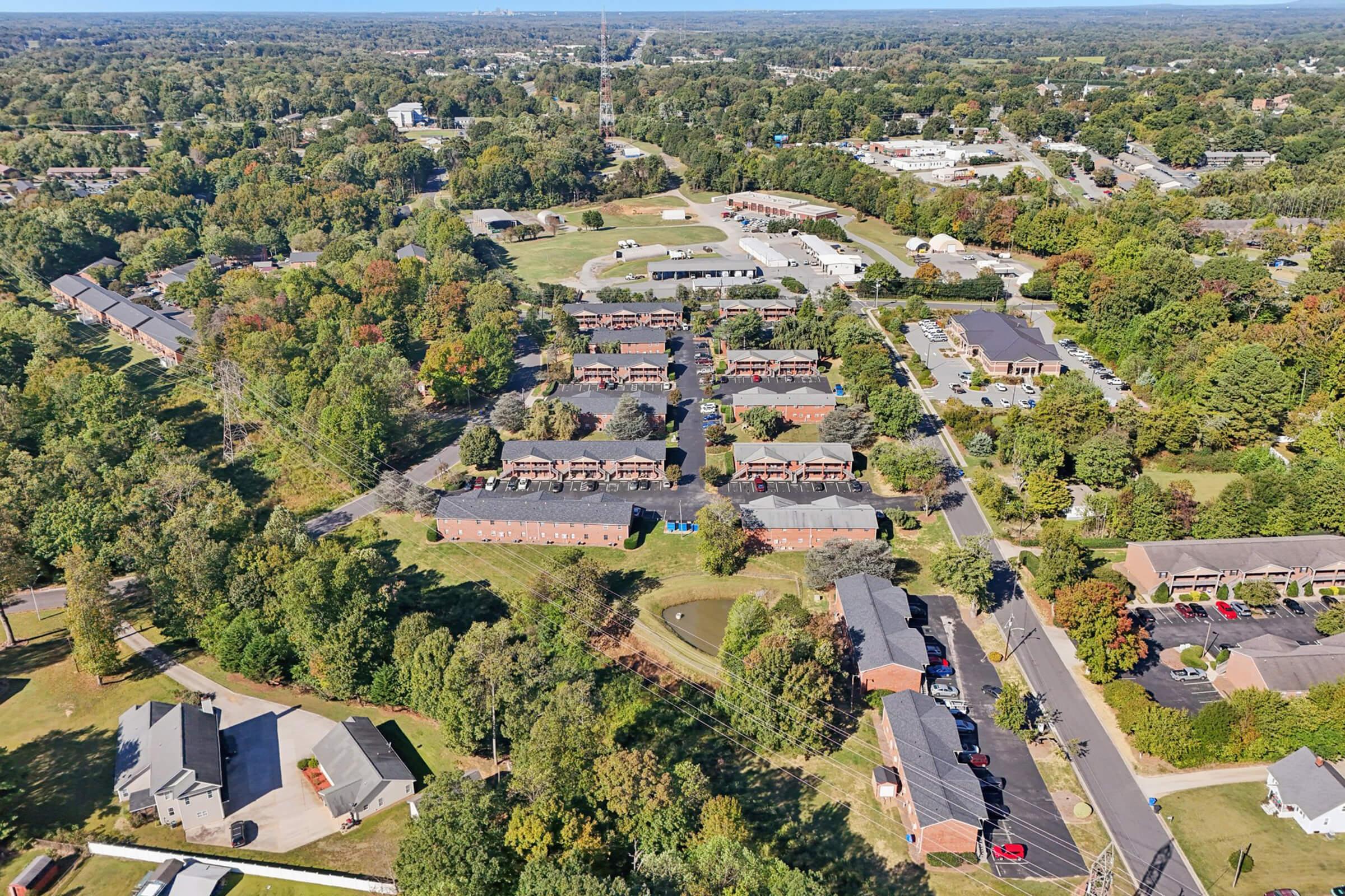 Aerial view of a suburban area featuring residential buildings, patches of greenery, and a golf course. The scene includes a mix of homes, commercial buildings, and trees under clear skies, showcasing a well-maintained neighborhood with various facilities nearby.