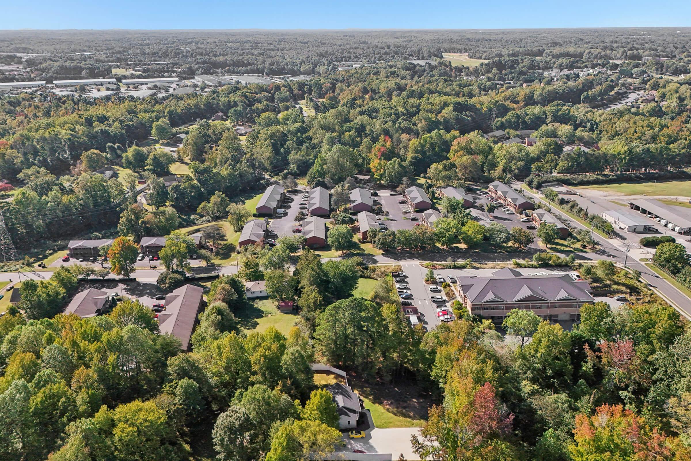 Aerial view of a suburban area featuring a cluster of residential buildings surrounded by trees. The landscape includes greenery, roads, and parking spaces, indicating a community or housing development amidst a natural setting. Clear skies and distant hills can be seen in the background.