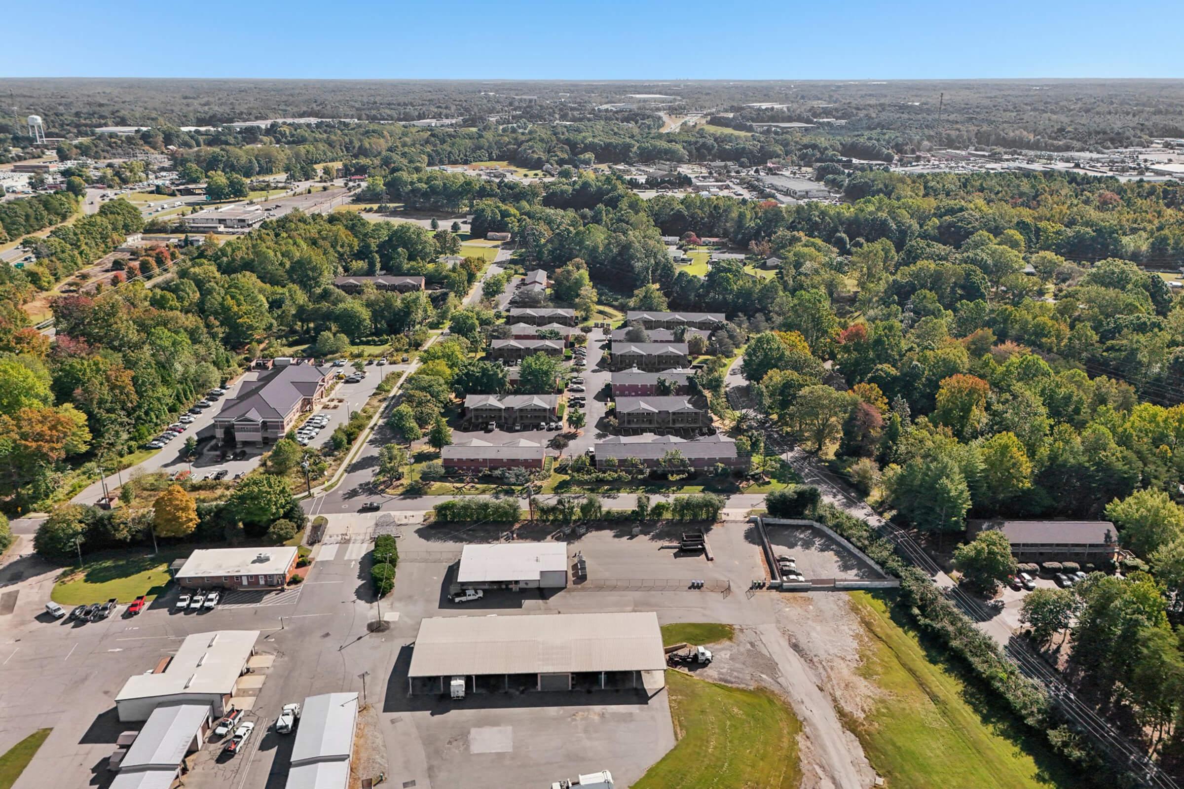Aerial view of a suburban area featuring clusters of residential buildings surrounded by trees. In the foreground, there are industrial structures and parking spaces. The background shows a highway and expansive green fields under a clear blue sky.