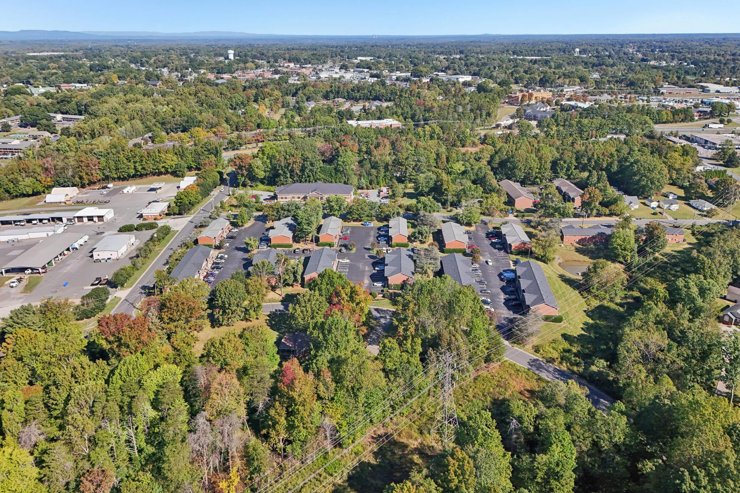 Aerial view of a suburban area featuring multiple residential buildings surrounded by trees, parking lots, and nearby commercial structures. The landscape shows a mix of greenery and developed land, with distant hills on the horizon under a clear blue sky.
