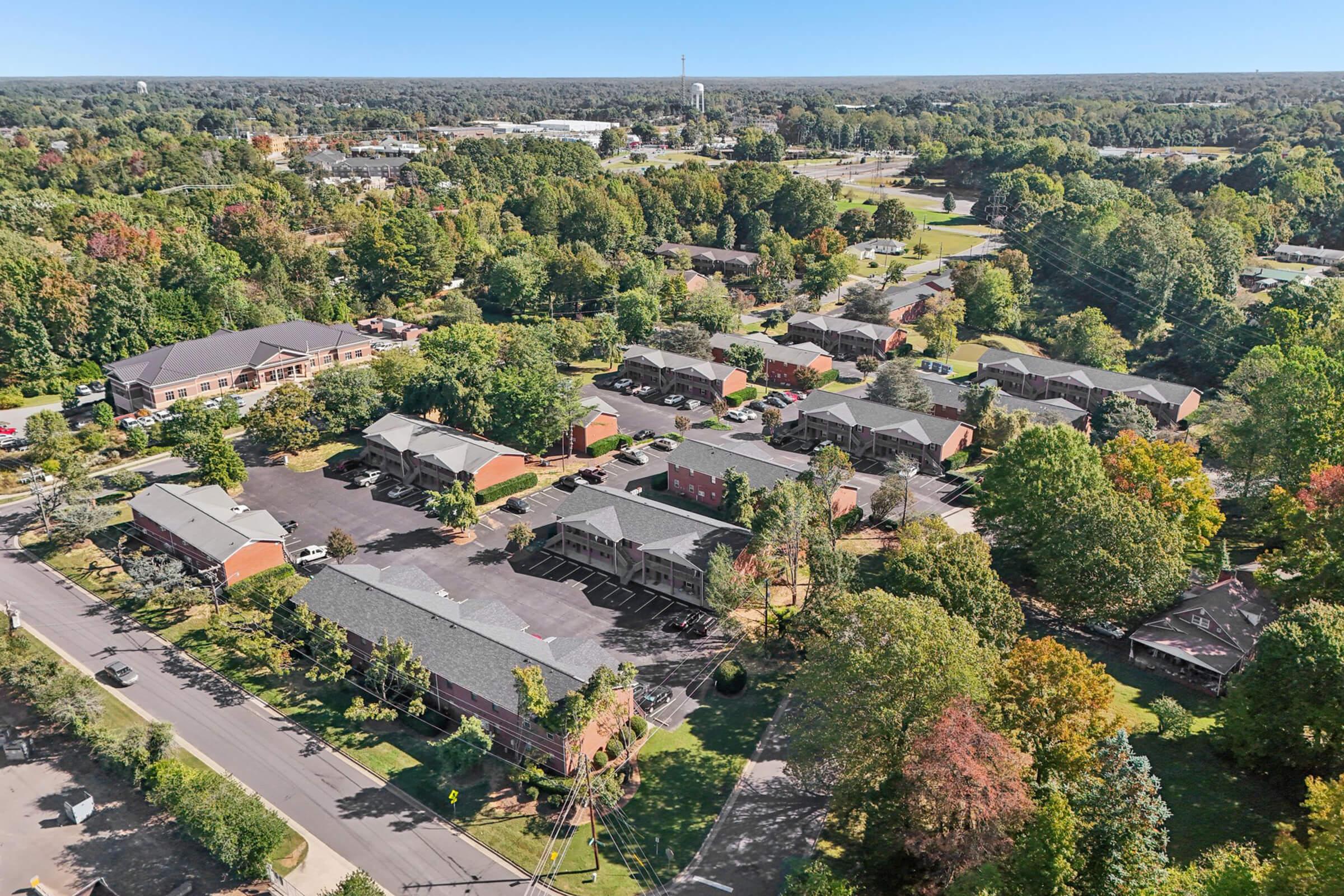 Aerial view of a suburban neighborhood featuring several buildings surrounded by trees with vibrant fall foliage, roads winding through the area, and a clear blue sky in the background. The landscape includes a mix of residential structures and green spaces, showcasing a tranquil community environment.