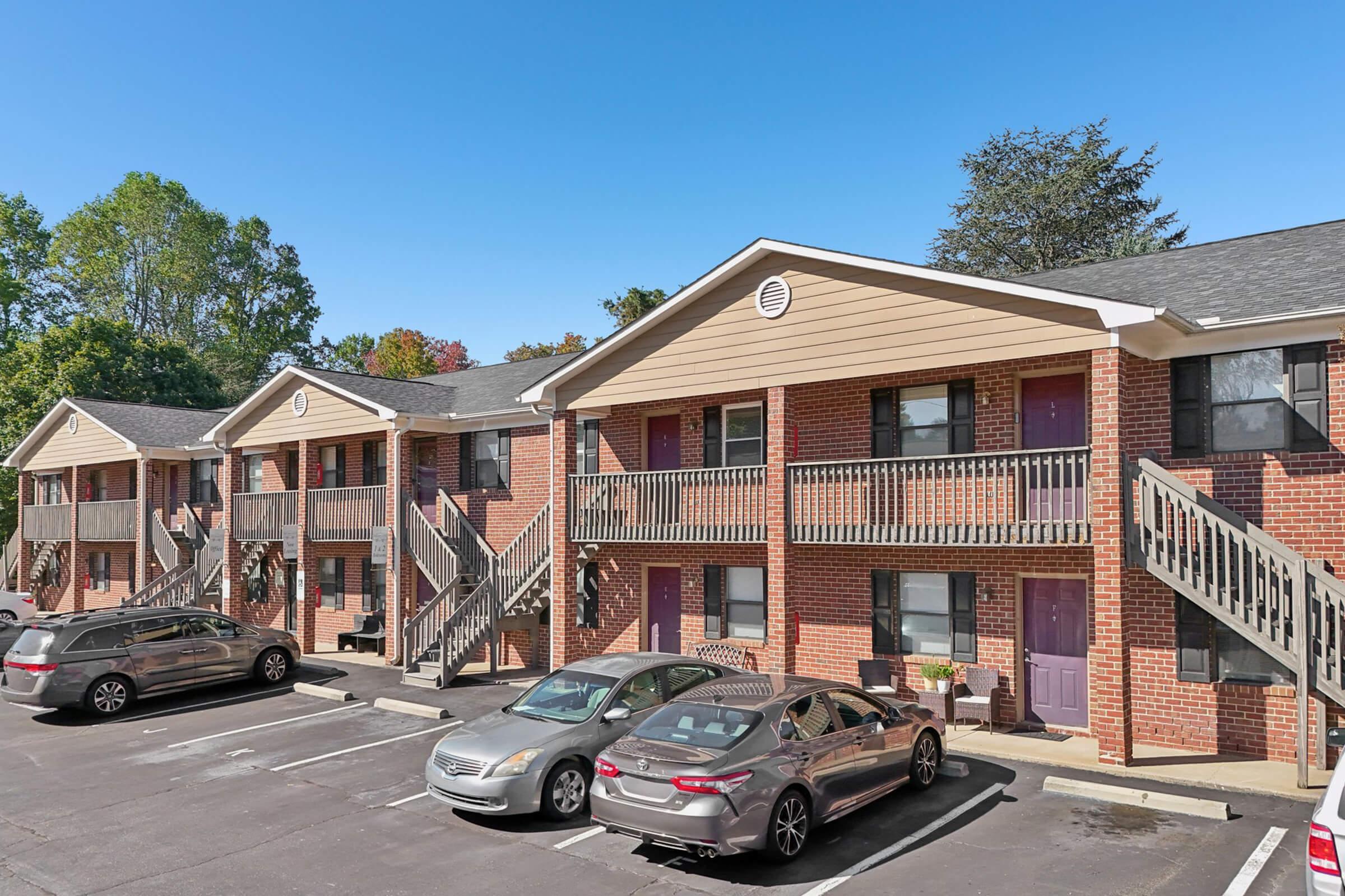 A row of apartment buildings featuring brick exteriors and wooden staircases leading to second-floor entrances. Several parked cars are visible in the foreground, and the scene is set against a clear blue sky with green trees in the background, indicating a pleasant residential area.
