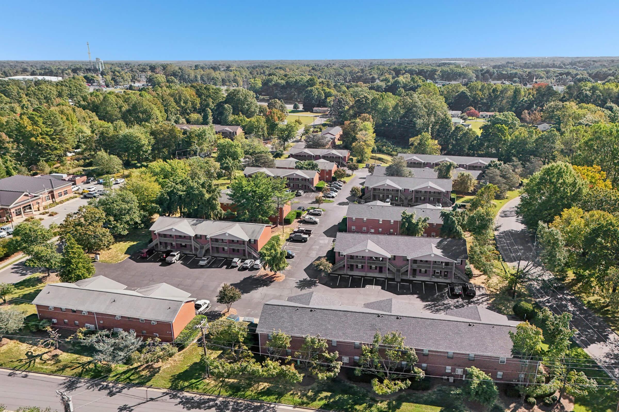Aerial view of a suburban apartment complex surrounded by trees and green spaces. The layout includes several buildings with parking areas, pathways, and lush landscaping. In the background, additional residential and commercial areas are visible, with a clear blue sky overhead.