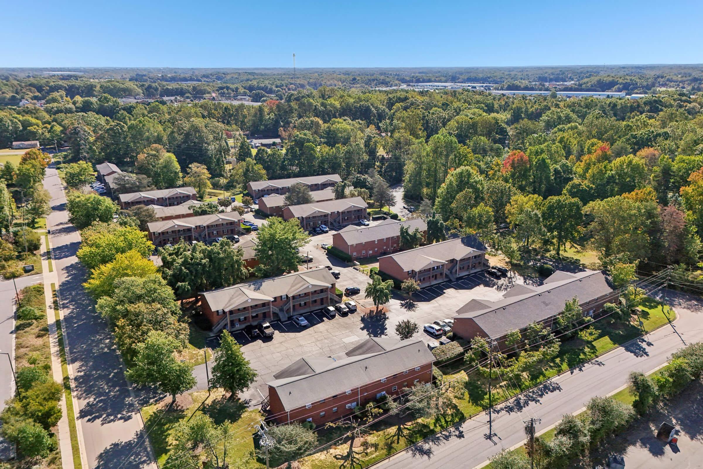 Aerial view of a residential neighborhood featuring several apartment buildings surrounded by trees with varying autumn colors. The area includes well-maintained lawns and parking spaces, with a road running alongside. In the distance, a winding river or road can be seen among the greenery.
