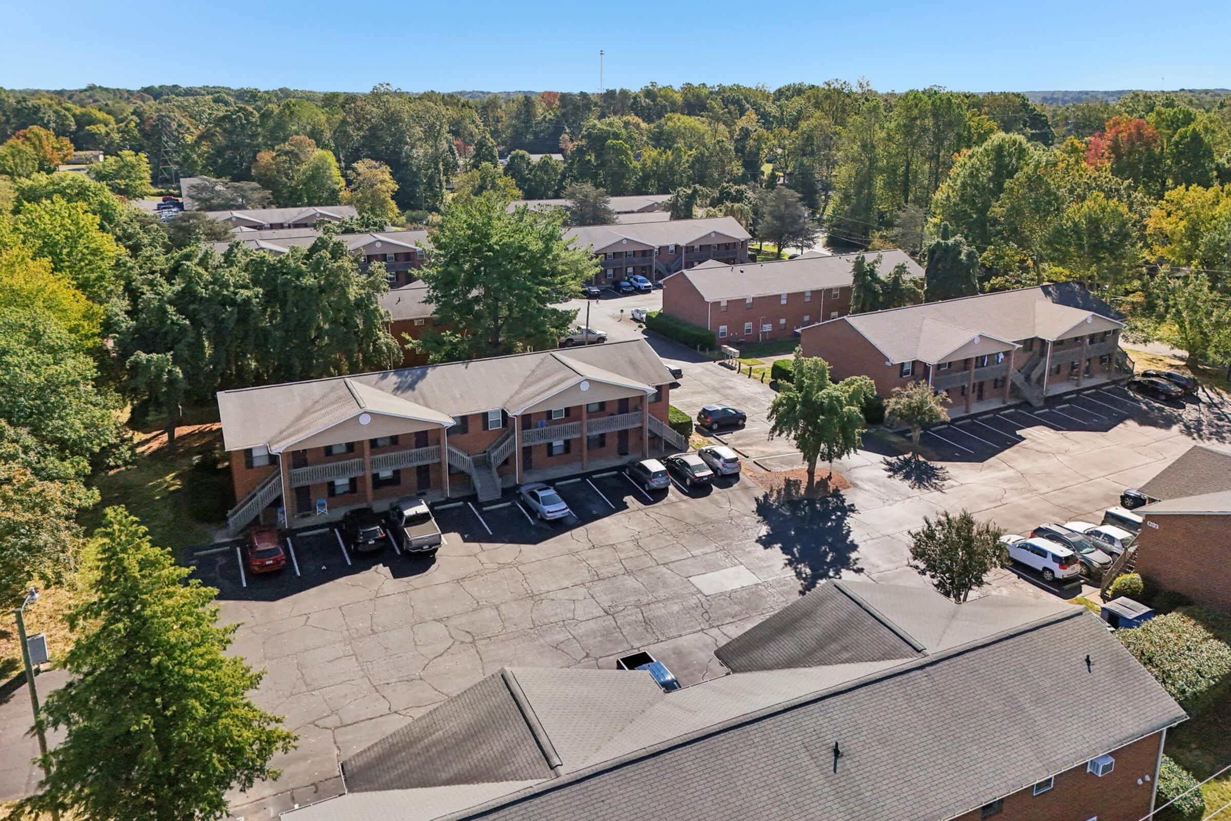 Aerial view of a residential complex featuring several buildings, surrounded by trees with autumn foliage. The parking area in front of the buildings has parked cars, and the landscape includes green grass and shrubs, creating a peaceful suburban atmosphere.