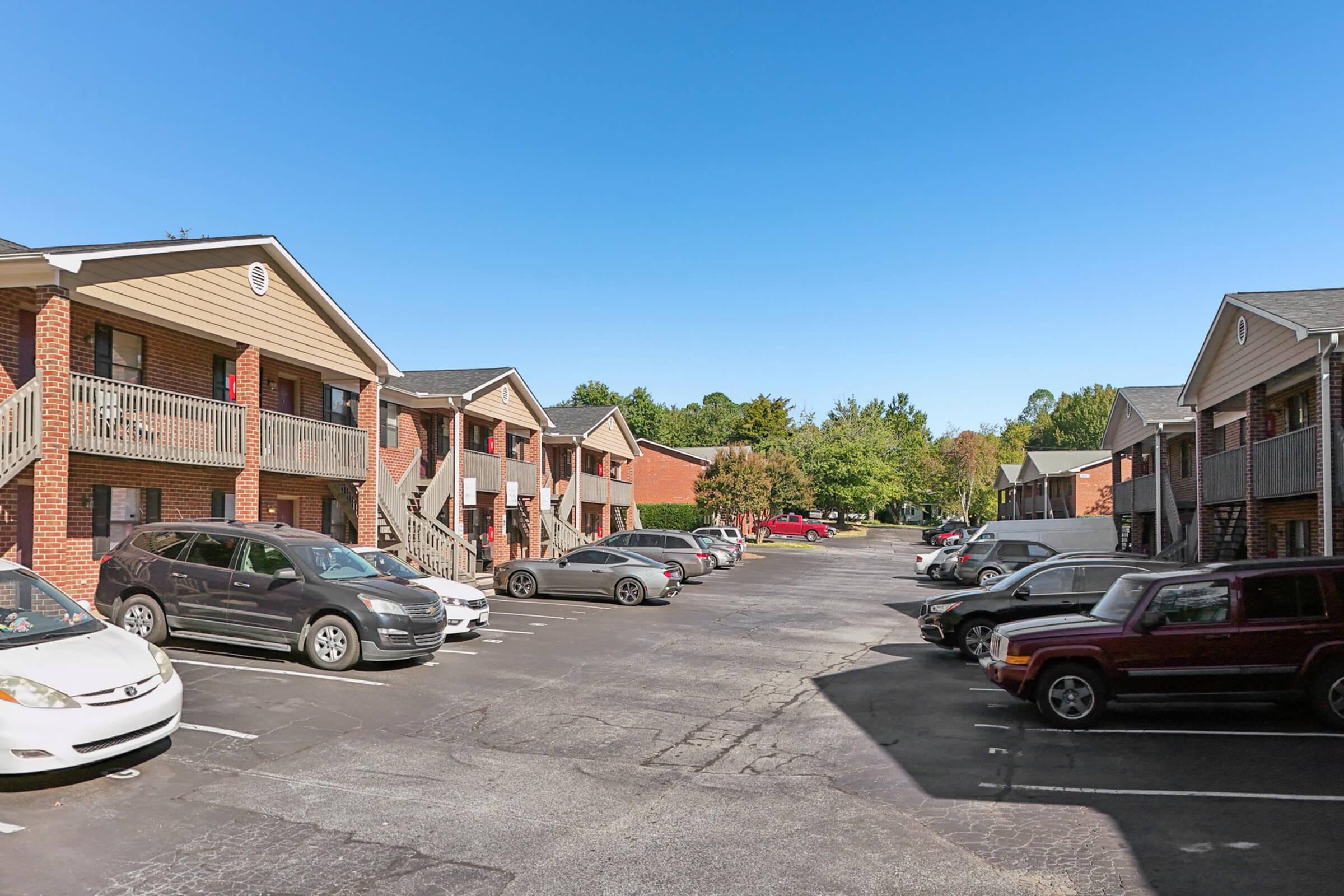 A view of a residential apartment complex featuring two-story buildings with balconies, surrounded by a parking lot filled with cars. The scene is set under a clear blue sky, with trees in the background providing a touch of greenery.