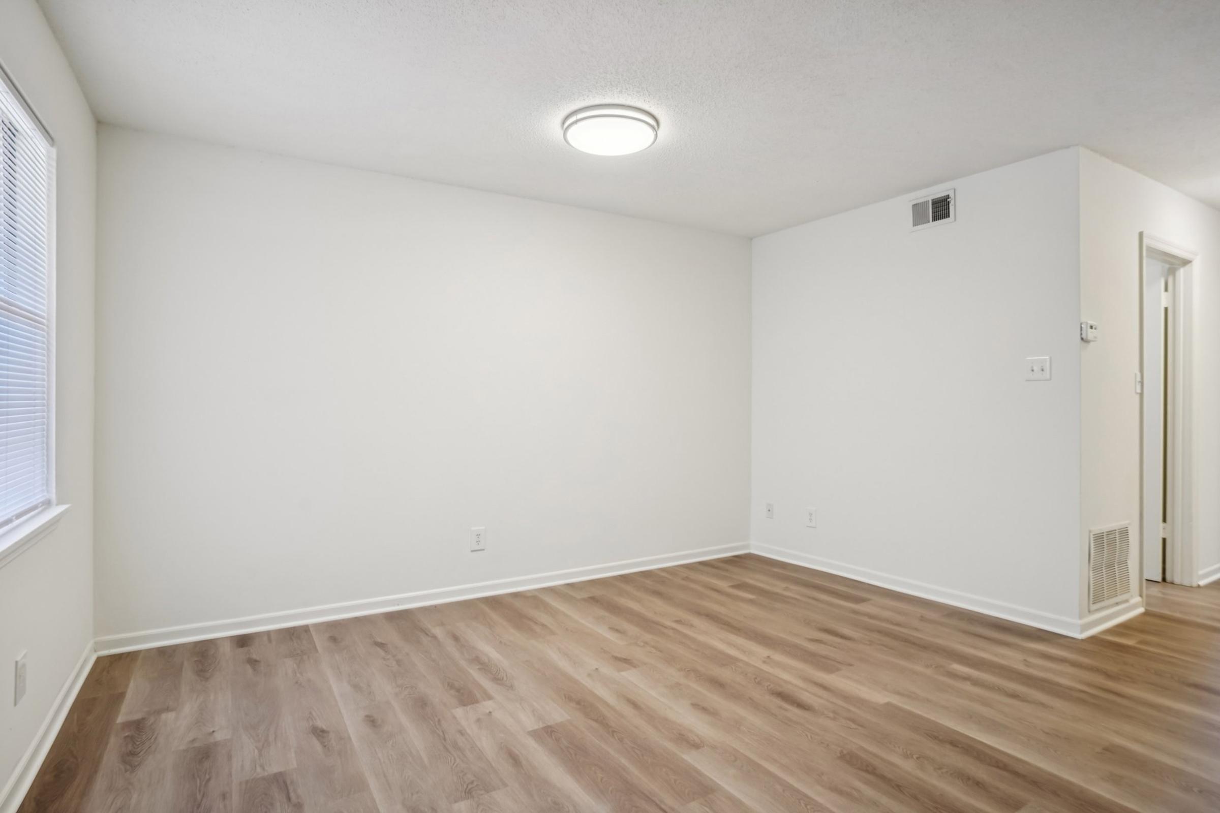 A spacious, empty room with light-colored walls and a smooth wooden floor. A ceiling light fixture is visible, and there are windows with natural light coming in. The room appears clean and uncluttered, with a door leading to another area at the back.