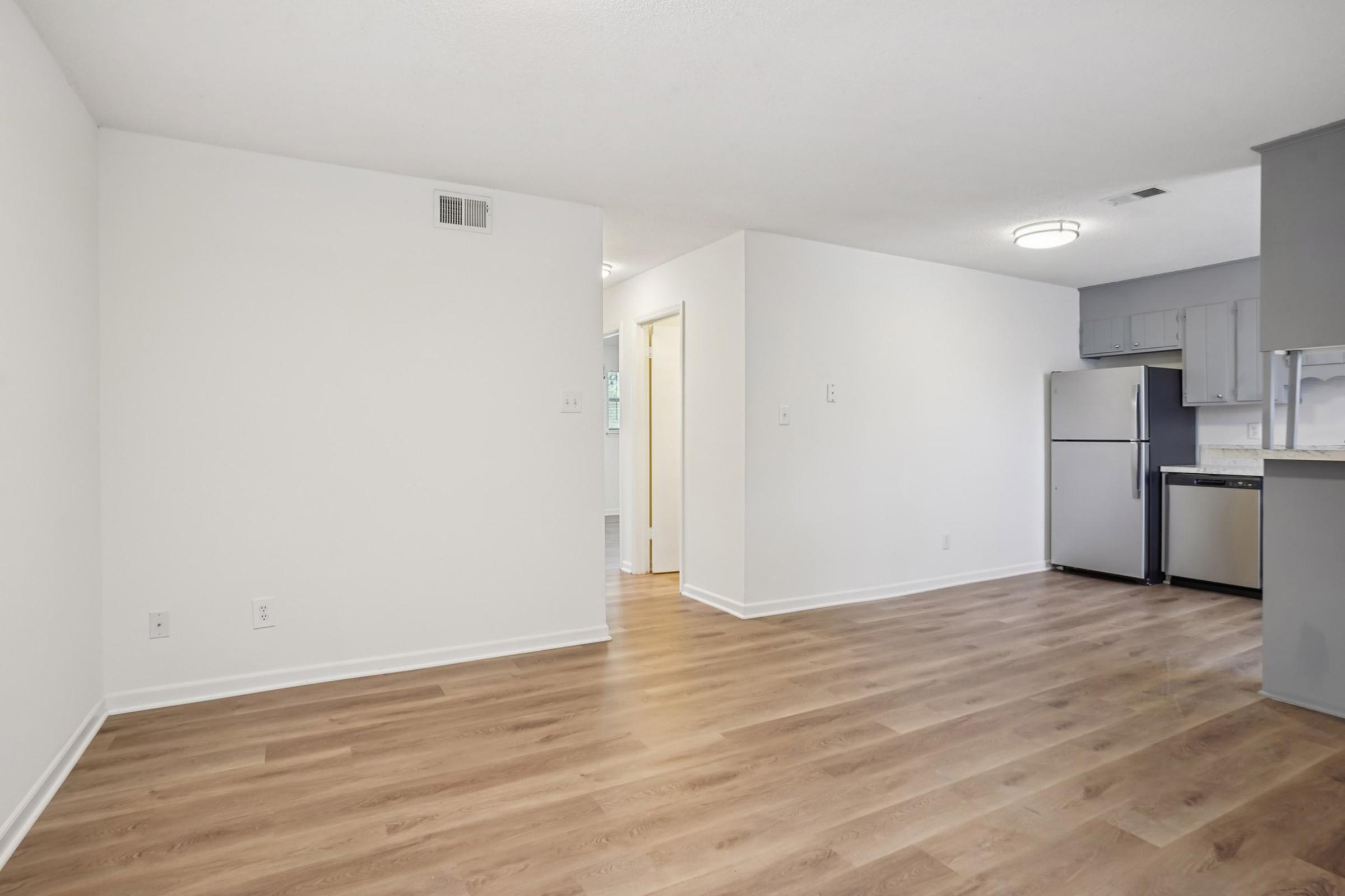 An interior view of a modern apartment featuring a spacious, empty living area with light-colored walls and light hardwood flooring. In the background, a small kitchen area with white cabinets, a stainless steel refrigerator, and a doorway leading to another room. Natural light illuminates the space.