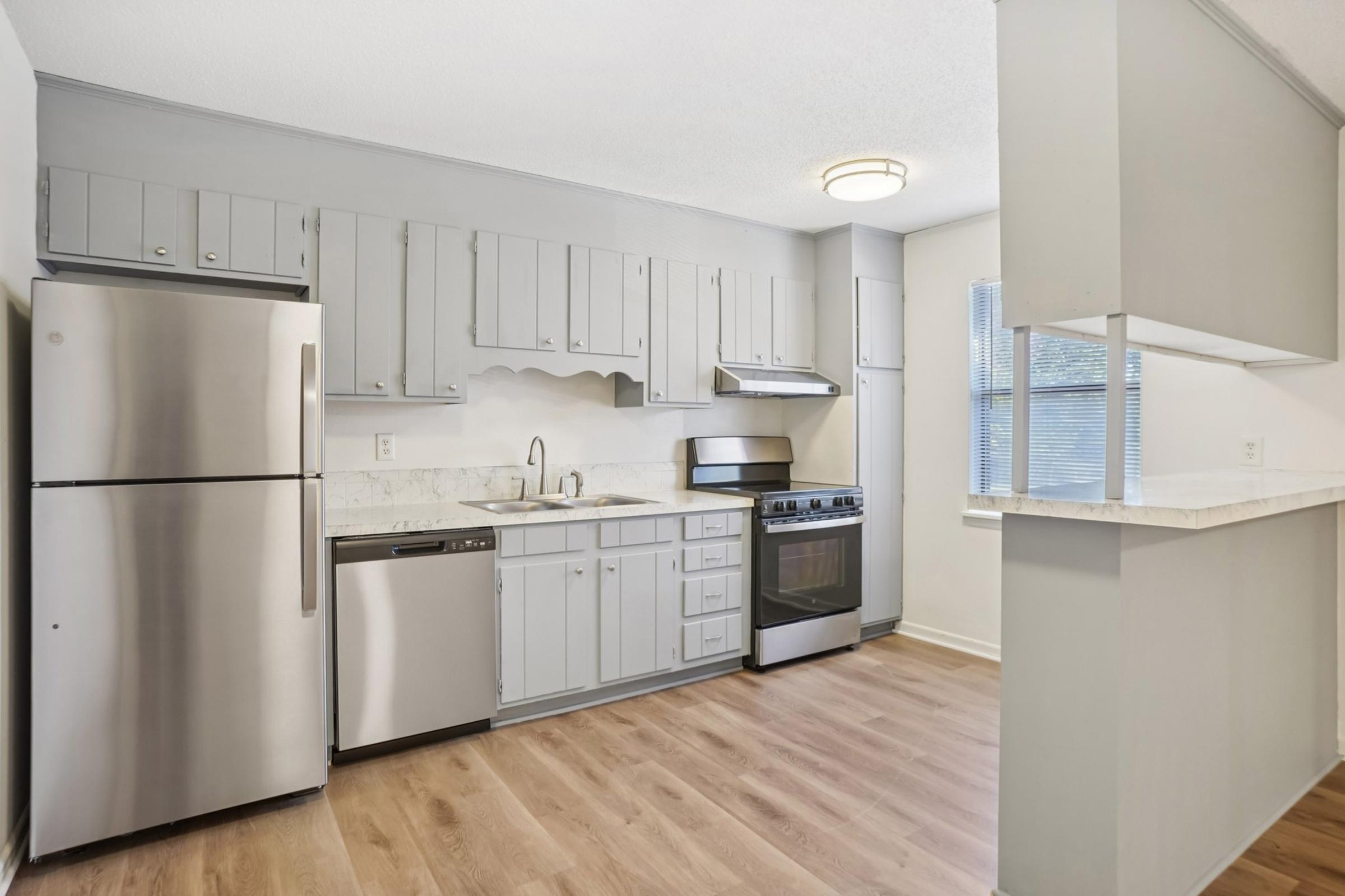 A modern kitchen featuring stainless steel appliances, including a refrigerator, dishwasher, and oven. The cabinets are light gray, and the countertops have a marble-like finish. Natural light enters through a window, and the flooring is a light wood laminate.