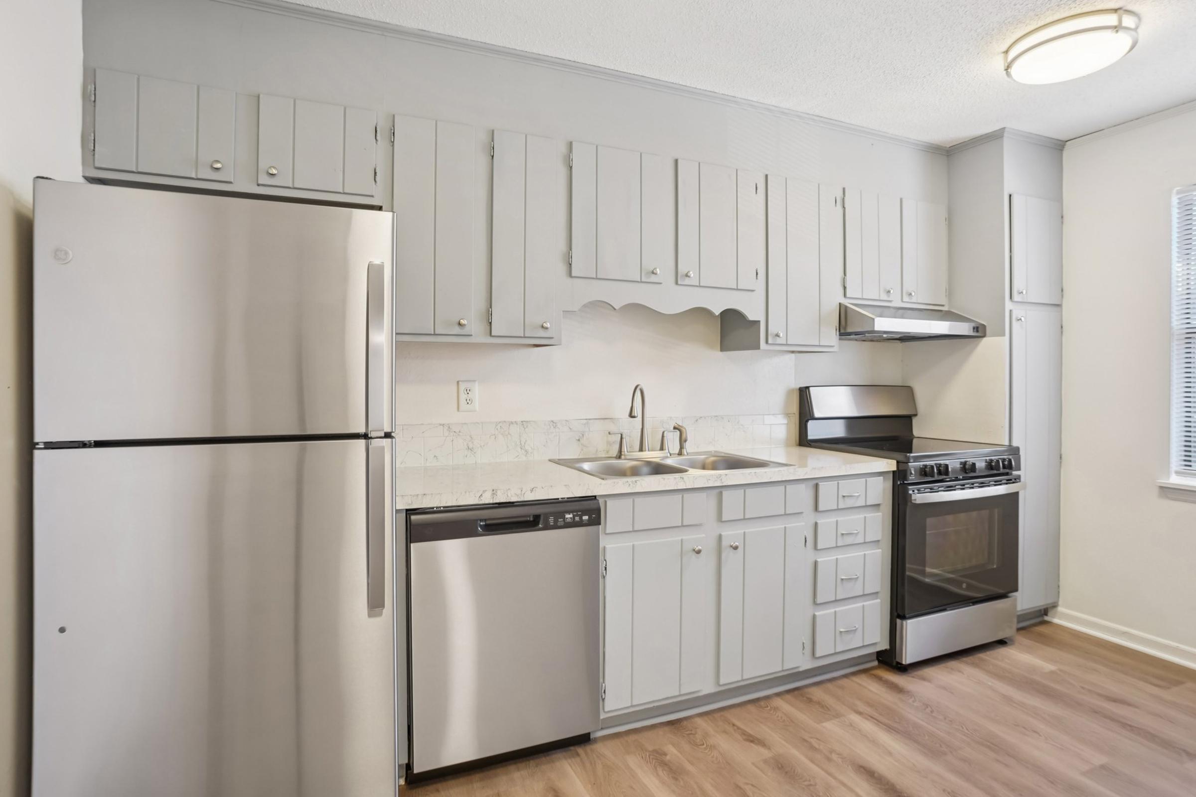 A modern kitchen featuring light gray cabinetry, stainless steel appliances including a refrigerator, dishwasher, and oven. The kitchen has a countertop with a double sink, and wood-like flooring. Natural light comes in through a window, enhancing the overall clean and contemporary look of the space.