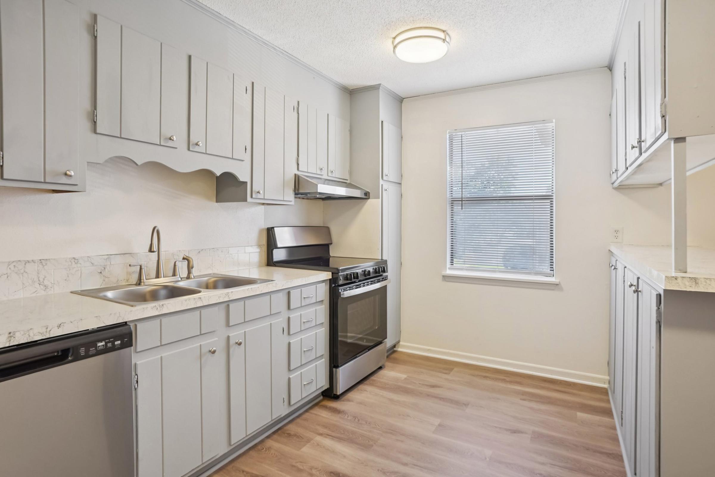 Modern kitchen featuring light gray cabinetry, stainless steel appliances including a stove and dishwasher, and a double sink. The countertop is a light marble design. There is a window providing natural light, and the floor has a warm wood finish.