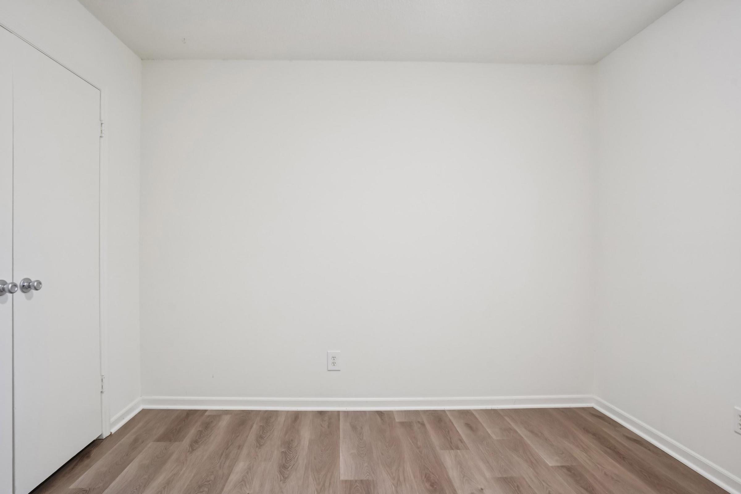 Empty room with light-colored walls and wooden flooring, featuring a closed white door on the left. The space is sparsely furnished, creating a minimalistic and clean aesthetic.