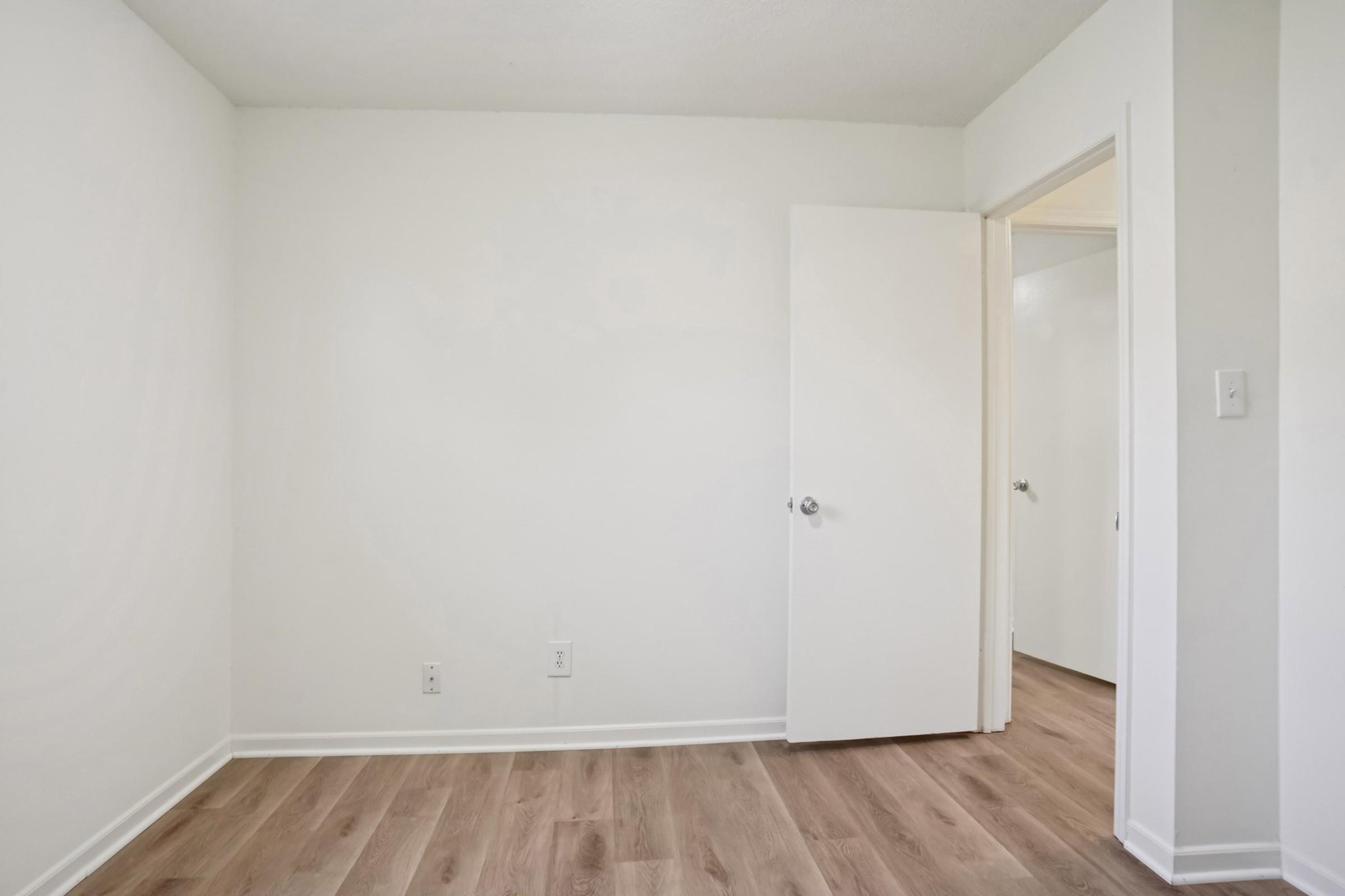 Empty room with light-colored walls and wooden flooring. There is a slightly ajar white door on the right leading to another room. Natural light illuminates the space, giving it a bright and airy feel. The room is unfurnished with no decorations, creating a minimalist appearance.