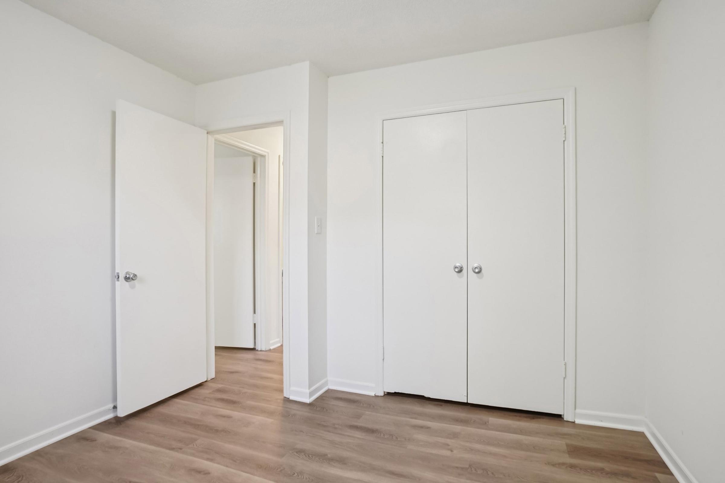 A bright, empty room with light-colored walls and wooden flooring. There are two white doors: one leading to an adjacent room and the other leading to a closet. The space appears clean and uncluttered, ideal for decorating or furnishing.
