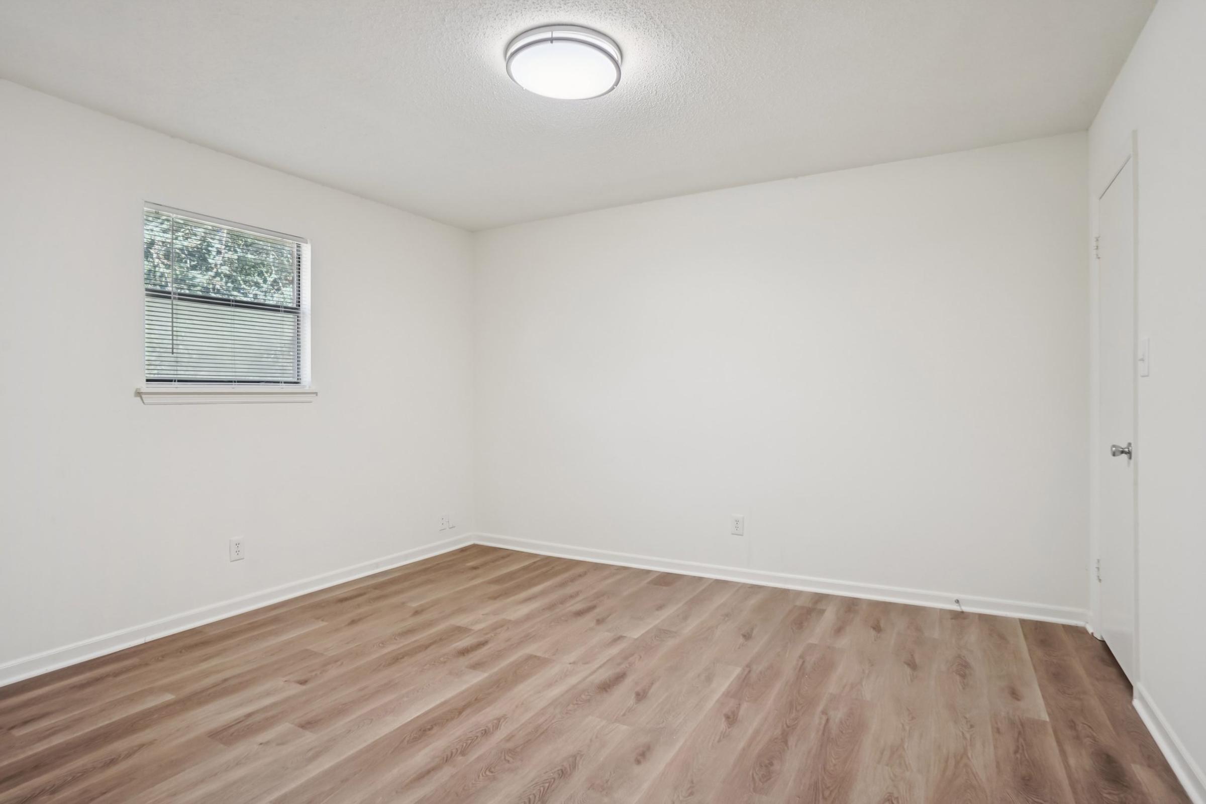 Empty room with light-colored walls, a textured ceiling, and a circular ceiling light. The floor is made of light brown wood laminate. There is a small window on the left side letting in natural light. The room appears spacious and freshly painted, with no furniture or decor present.