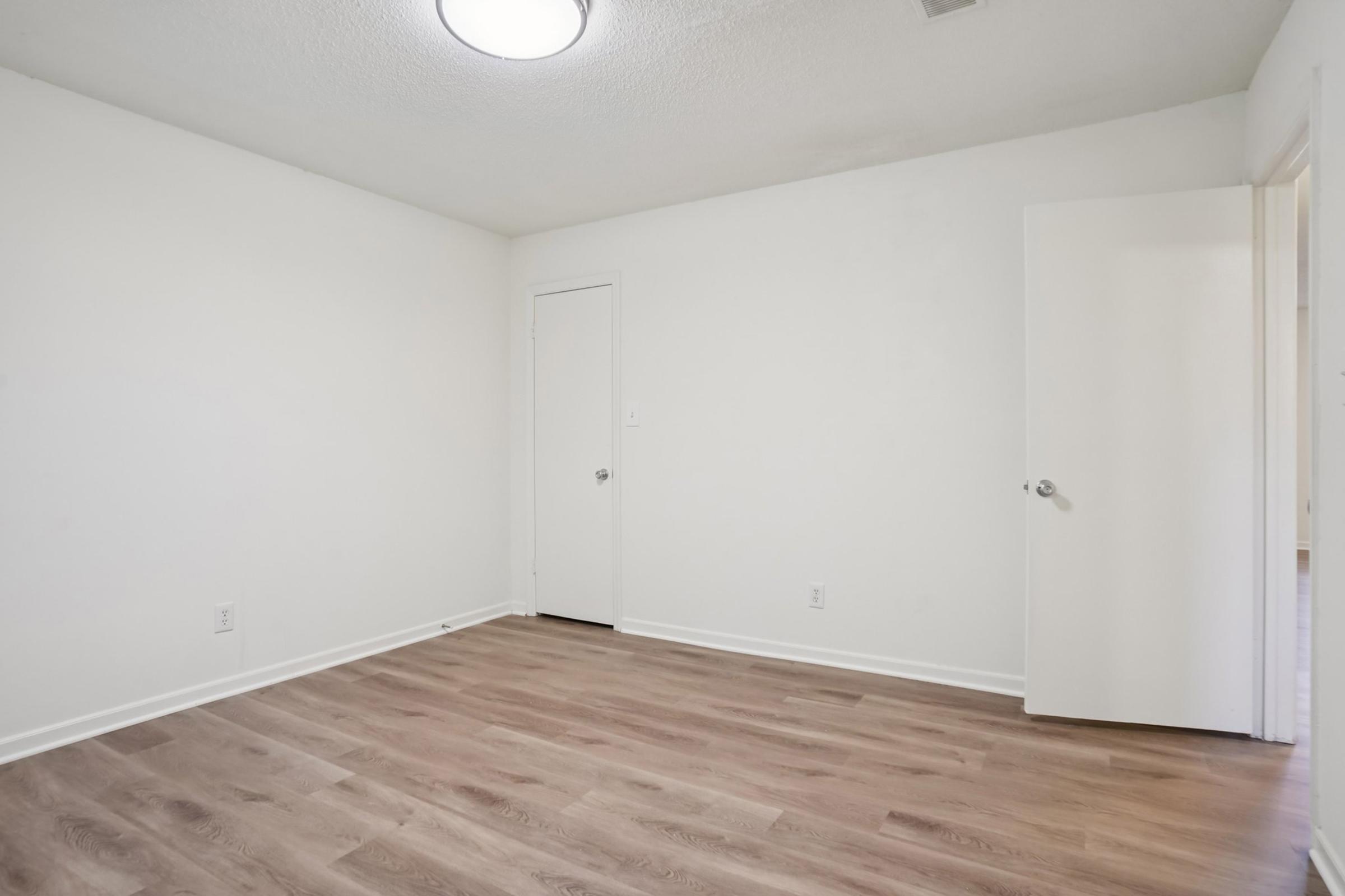 A clean, empty room with light-colored walls and a wooden floor. There is a ceiling light fixture, a closed door on the left, and an open door leading to another space on the right. The room appears well-lit and spacious, suitable for various uses.
