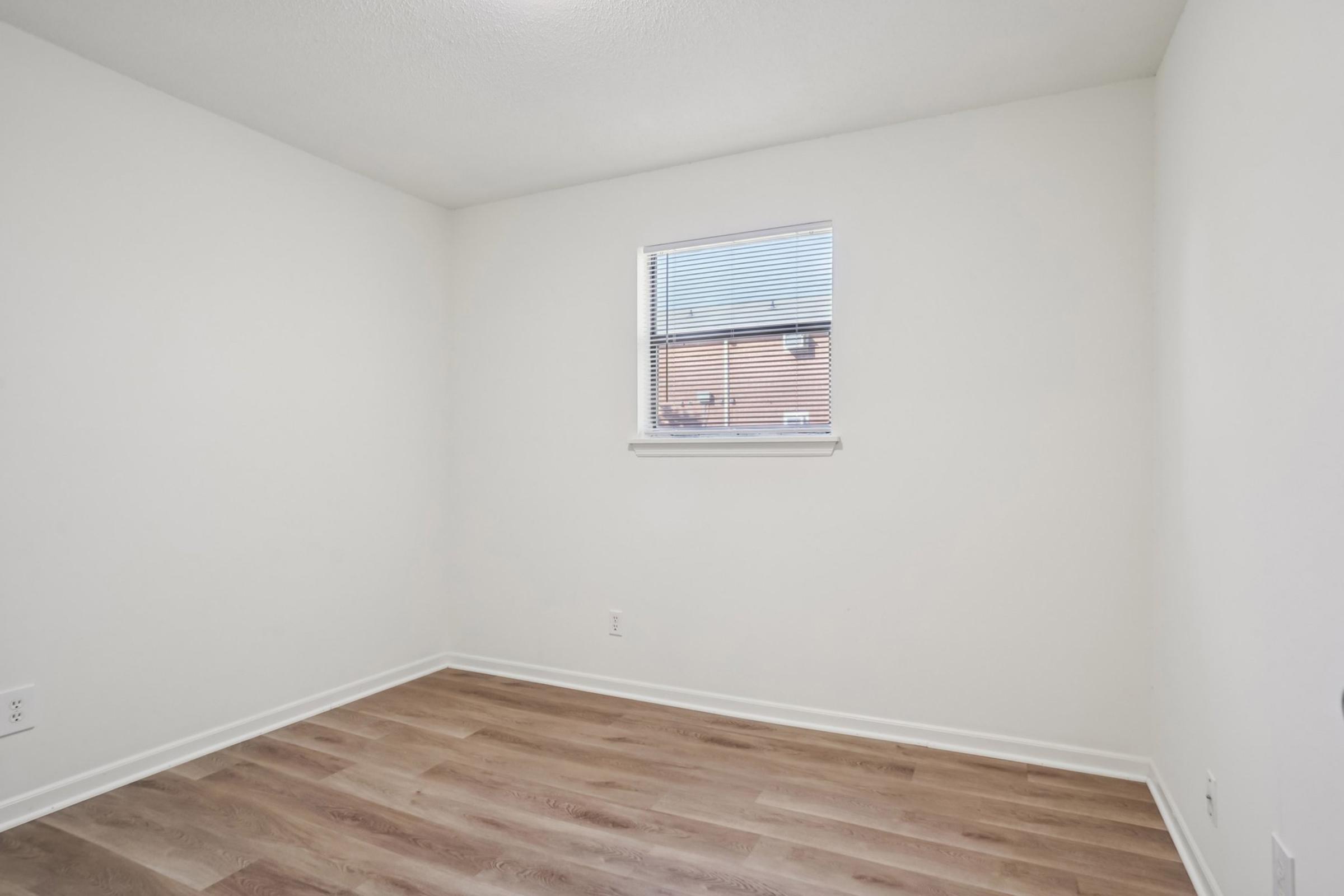 Empty room with light-colored walls and a wooden floor. A small window with blinds allows natural light to enter. The space is minimalistic and uncluttered, suitable for various uses such as an office or bedroom.