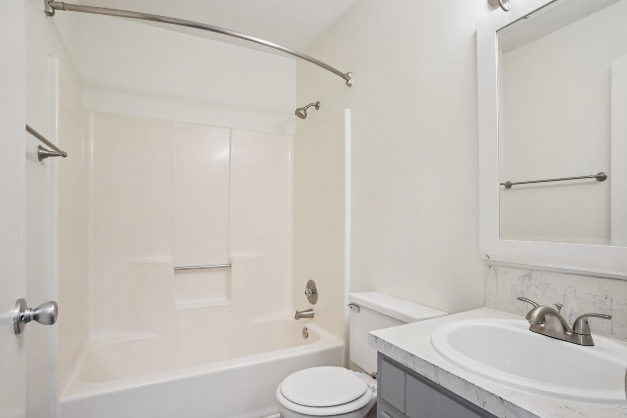A clean, modern bathroom featuring a white bathtub with a curved shower rod, a toilet, and a sink with a marble countertop. The walls are light-colored, and there is a simple mirror above the sink, contributing to a bright and airy atmosphere.