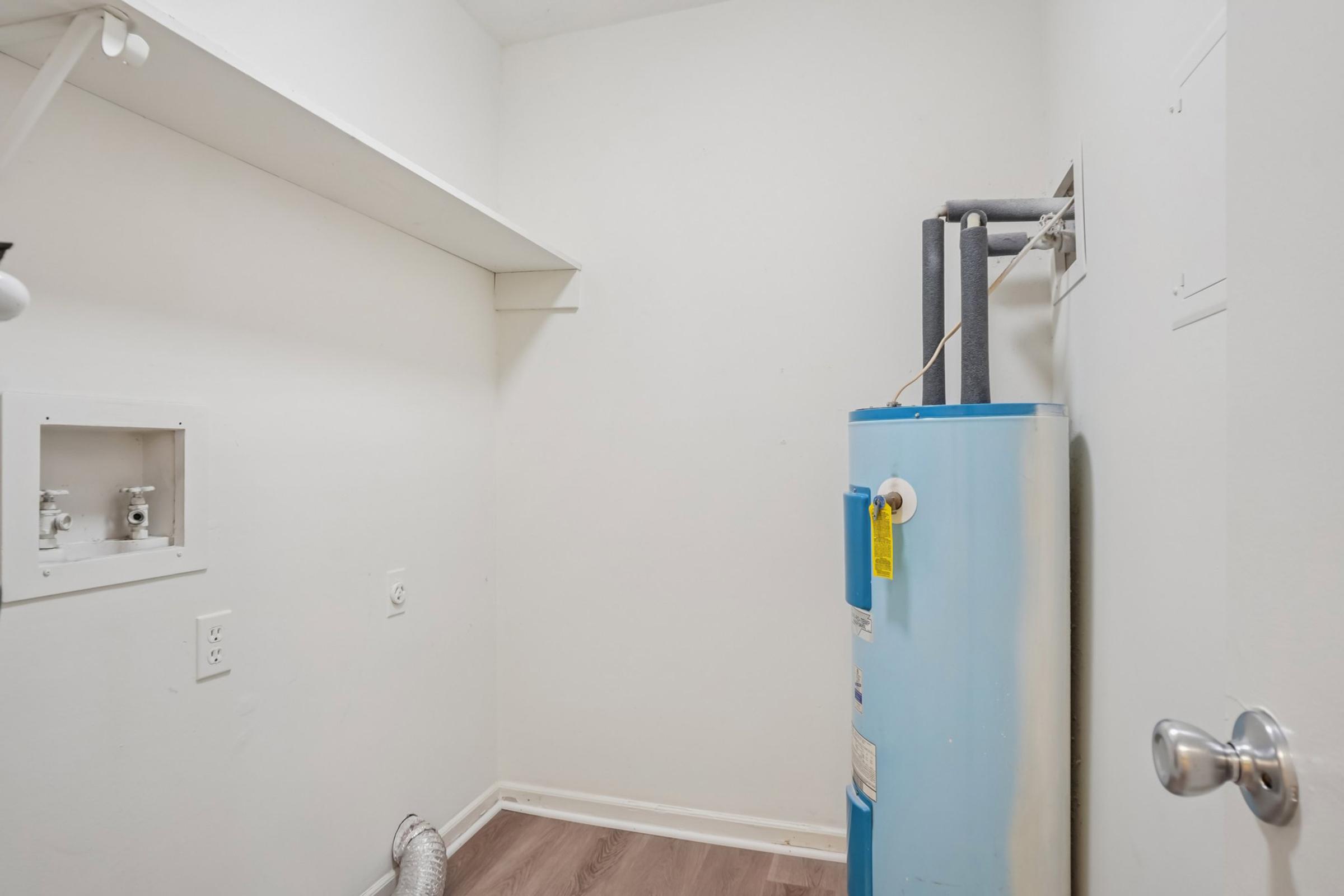A small, empty laundry room featuring a blue water heater against a white wall. There is a shelf above, and the floor is covered with light wood laminate. The room has a door with a silver handle and a vent. The overall space appears clean and uncluttered, suitable for laundry appliances.