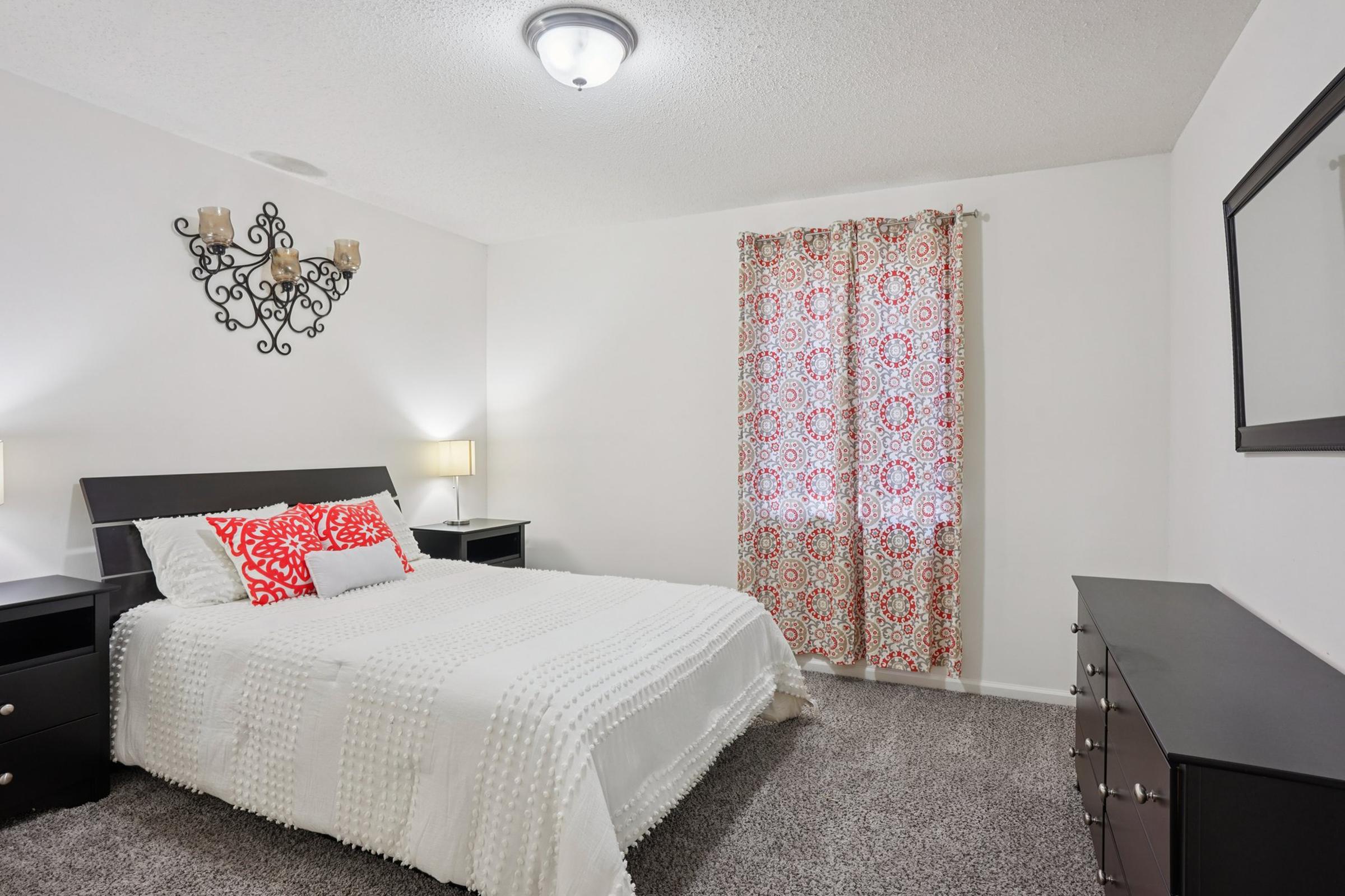 Bright bedroom featuring a queen-sized bed with a white blanket and red accent pillows. There are two bedside lamps on black nightstands, a dresser, and a decorative wall sconce. Large window covered with floral-patterned curtains allows natural light, while the gray carpet adds warmth to the space.