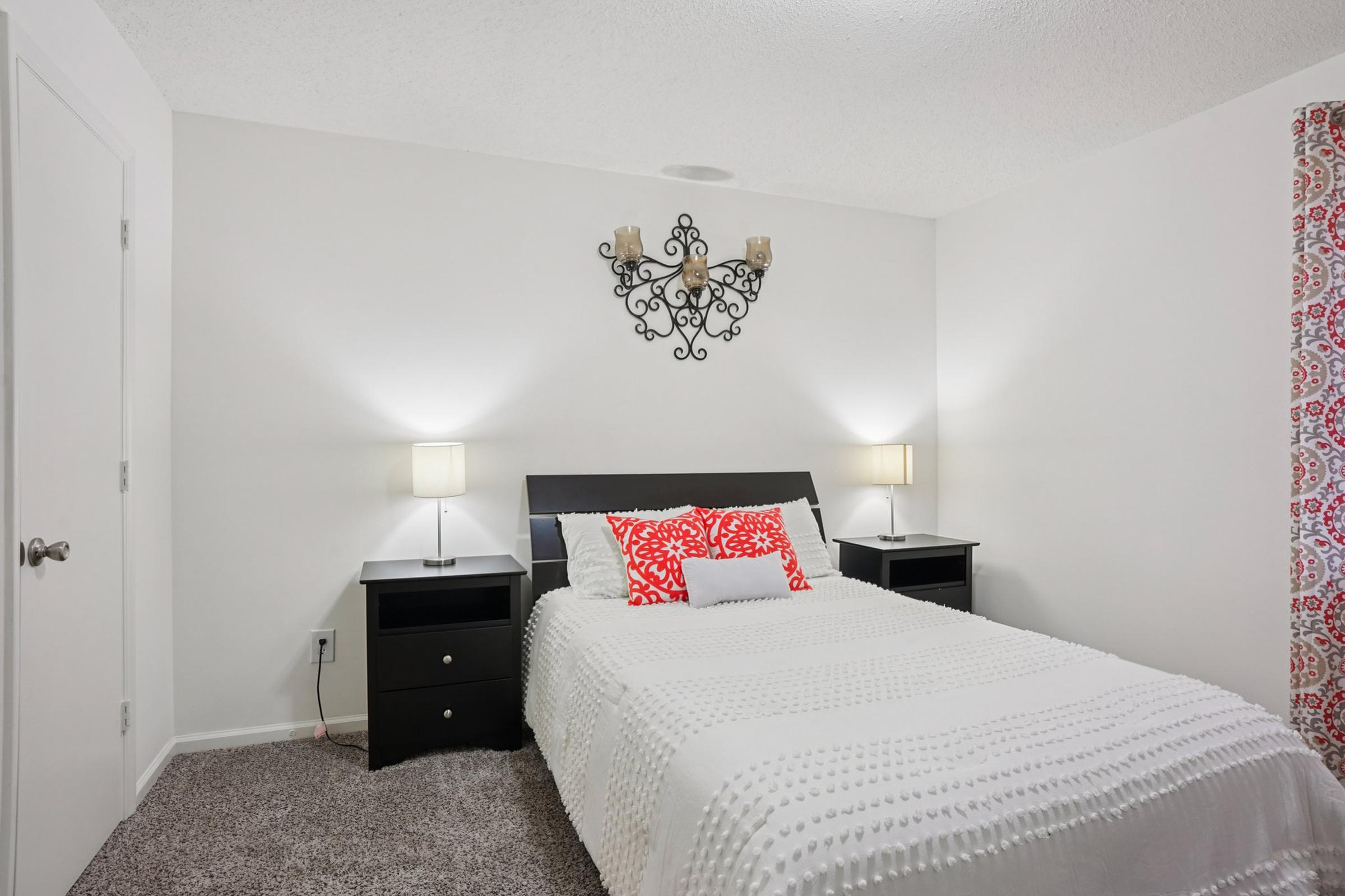 A cozy bedroom featuring a white bedspread with decorative pillows, two bedside tables with lamps, and a wall-mounted chandelier. The room has light-colored walls, carpeted flooring, and a patterned window curtain, creating a warm and inviting atmosphere.