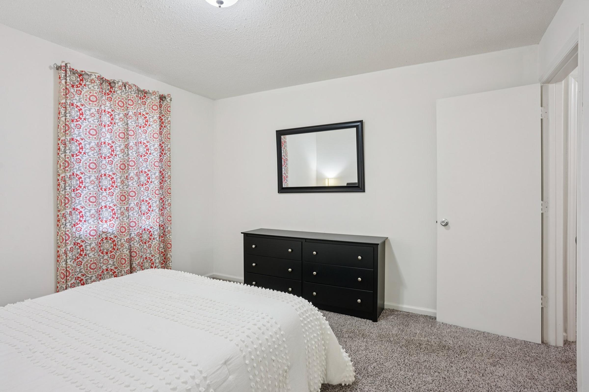 A bright bedroom featuring light-colored walls and carpet, a queen-sized bed with a textured white blanket, black dresser, a large mirror on the wall, and colorful patterned curtains. The room has a warm and inviting atmosphere with natural light coming in.