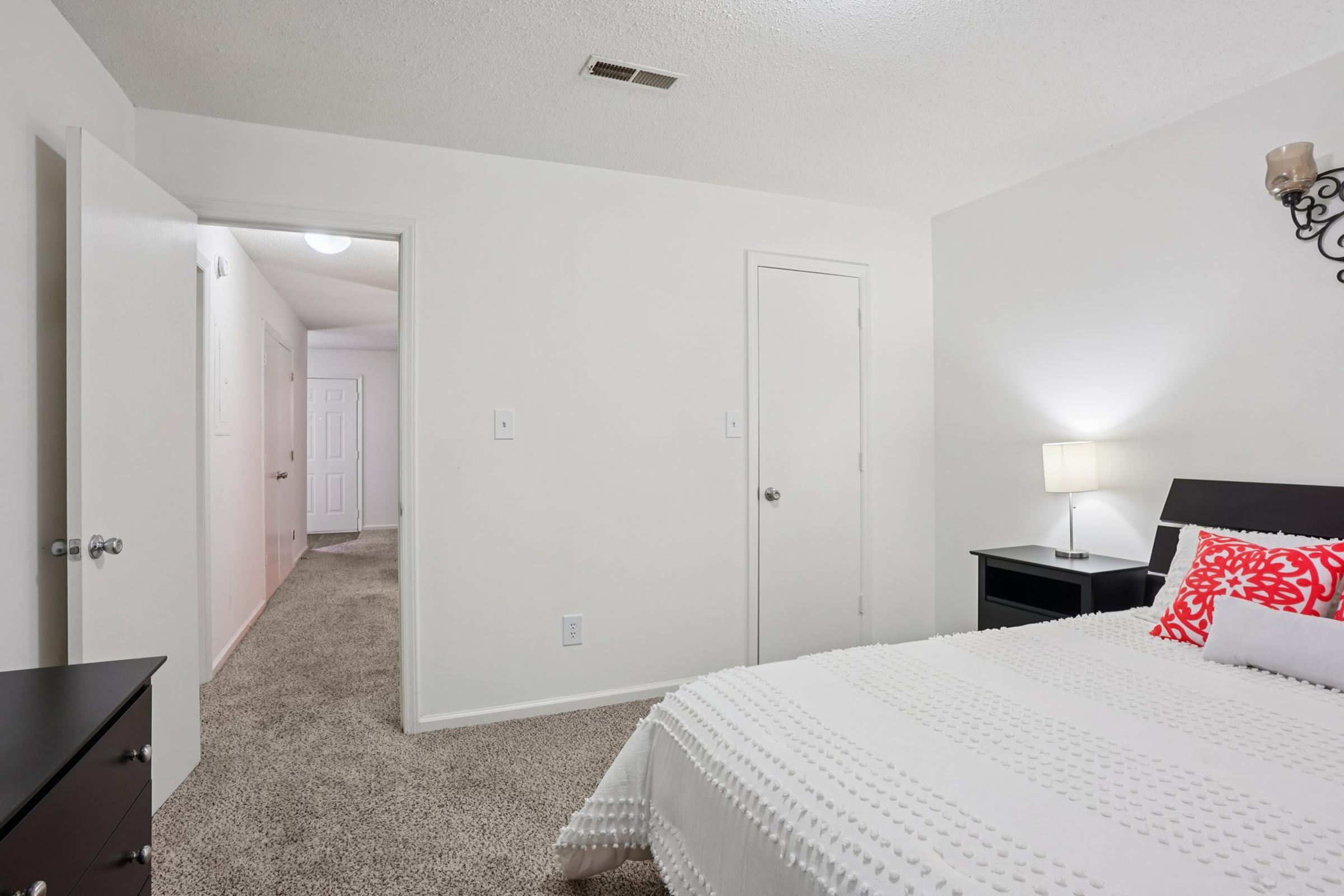 A bright, clean bedroom featuring a bed with white bedding and a red accent pillow. There's a bedside table with a lamp, and a large, neutral-colored carpet. A doorway leads to a hallway with closed doors on either side, enhancing the room's spacious feel. The walls are painted white, creating a minimalist atmosphere.
