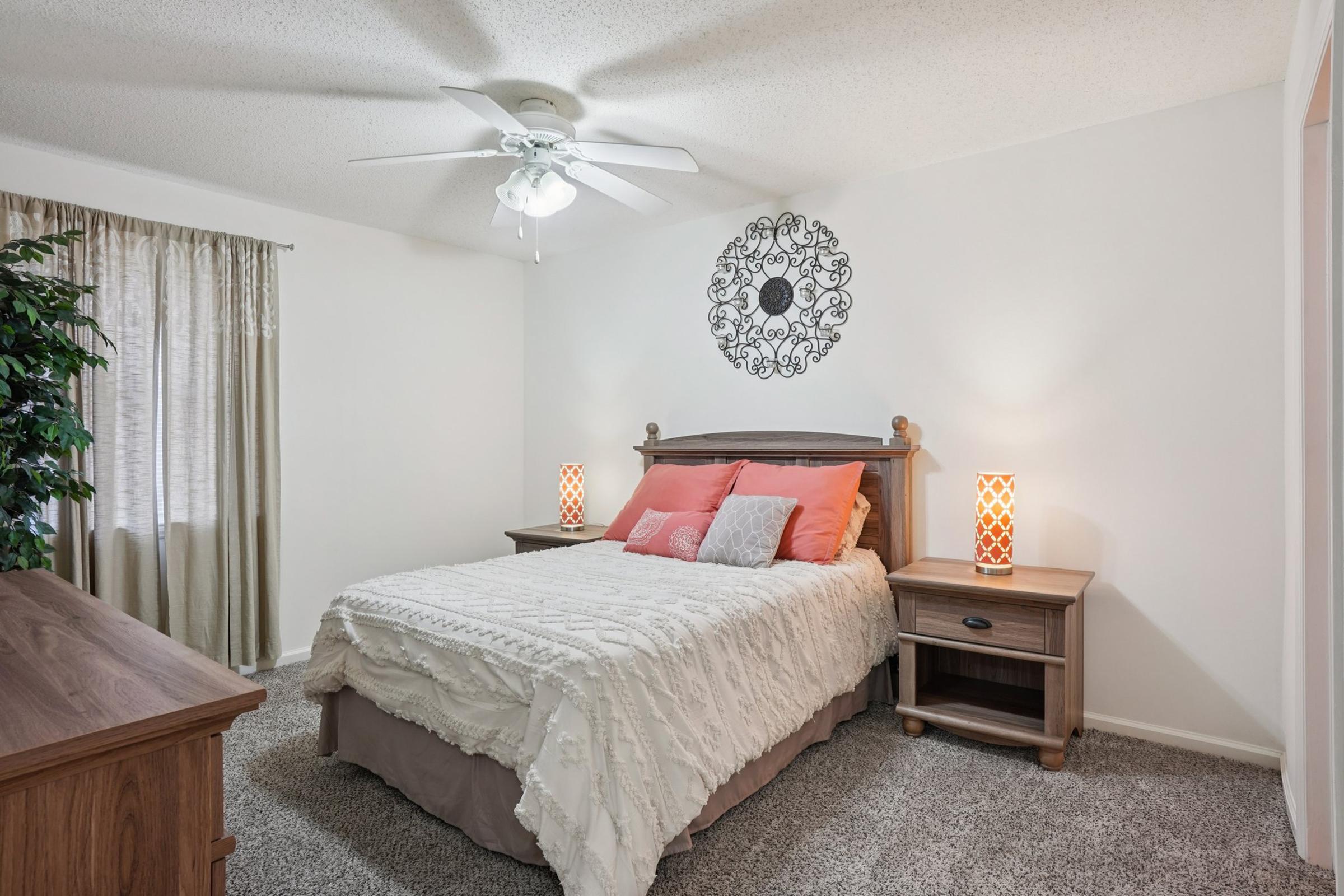 A cozy bedroom featuring a queen-sized bed with white bedding and decorative orange pillows. There are two stylish lamps on nightstands, a wooden dresser, and a large window with sheer curtains. A decorative wall piece adds character to the space, and a potted plant provides a touch of greenery.