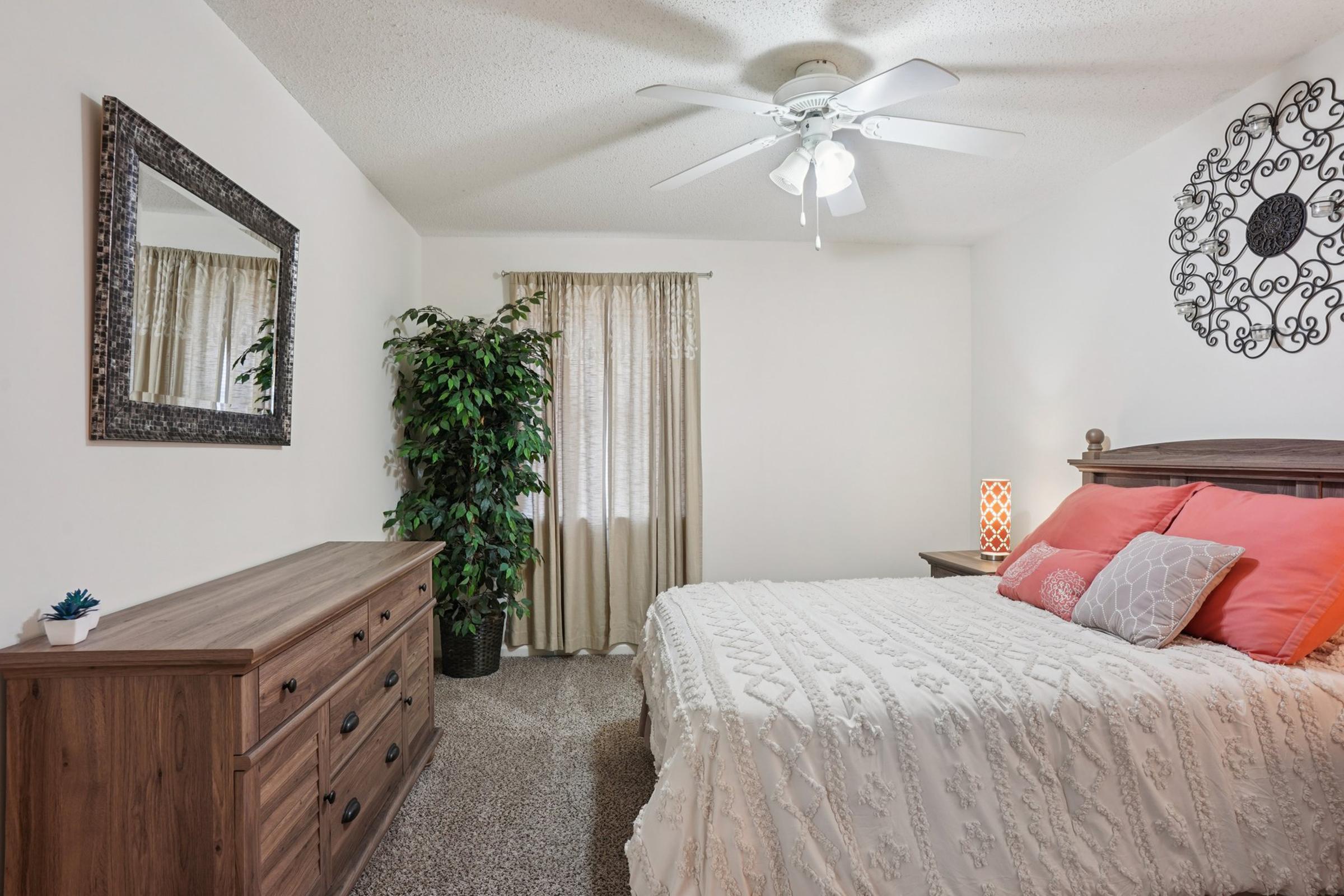 A cozy bedroom featuring a queen-sized bed with decorative pillows, a wooden dresser, a mirror, and a potted plant. The walls are light-colored, and there's a ceiling fan above. Natural light filters in through a curtained window, creating a warm and inviting atmosphere.