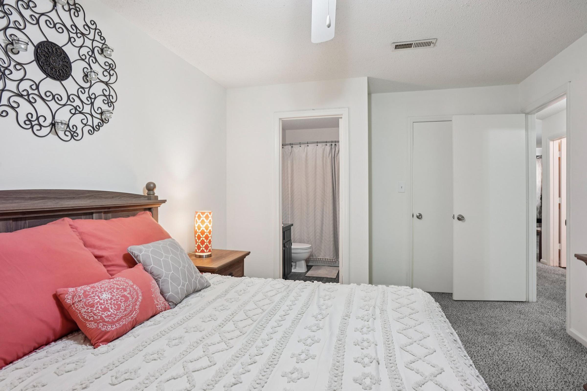 A cozy bedroom featuring a large bed with pink and gray decorative pillows. A stylish wall decor piece hangs above the bed. To the right, there's an open closet door and a bathroom entrance with a shower curtain visible. The space is well-lit and has a soft, inviting atmosphere.