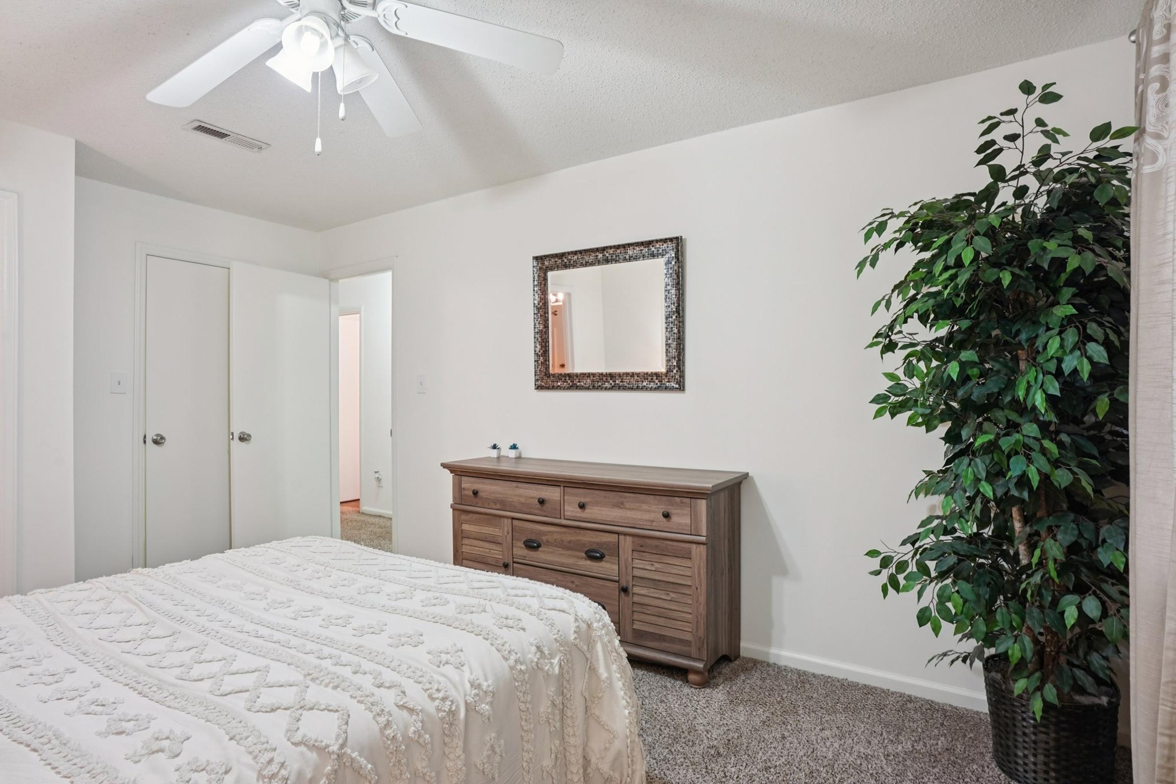A cozy bedroom featuring a light-colored bedspread on a bed, a wooden dresser with decorative items, a mirror on the wall, and a large artificial plant in the corner. The walls are painted white, and there are two doorways leading to other rooms, with carpeted flooring adding warmth to the space.