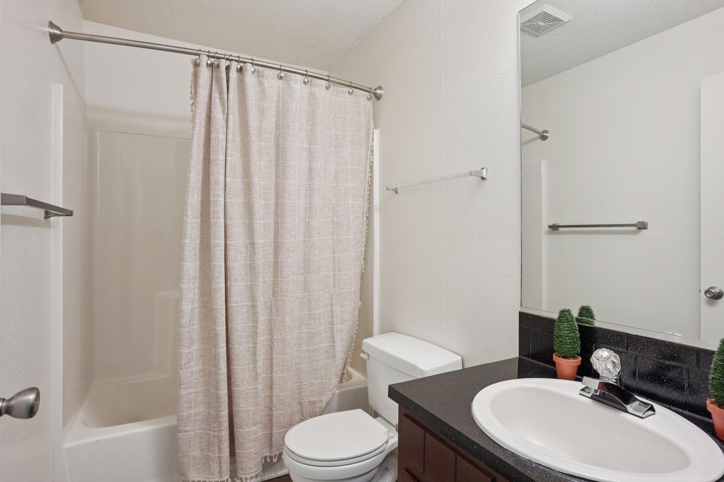 A clean bathroom featuring a bathtub with a shower curtain, a toilet, and a sink with a dark countertop. Green potted plants are placed on the countertop, adding a touch of decor. The walls are light-colored, and there are metal towel bars on the wall.