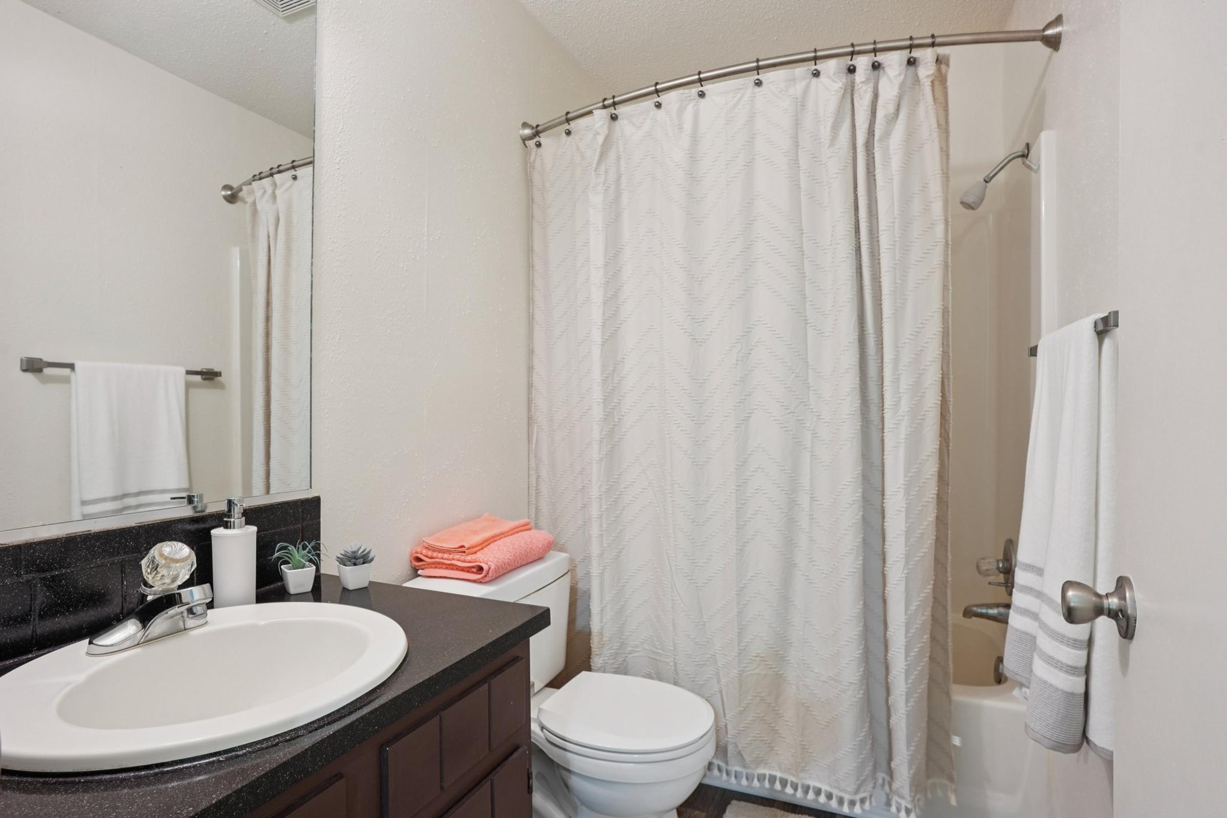 A clean and modern bathroom featuring a round sink with a sleek faucet, a dark countertop, a white shower curtain with a geometric pattern, and neatly arranged towels in coral and white. The space is illuminated with neutral walls and includes a small potted plant for a touch of decor.
