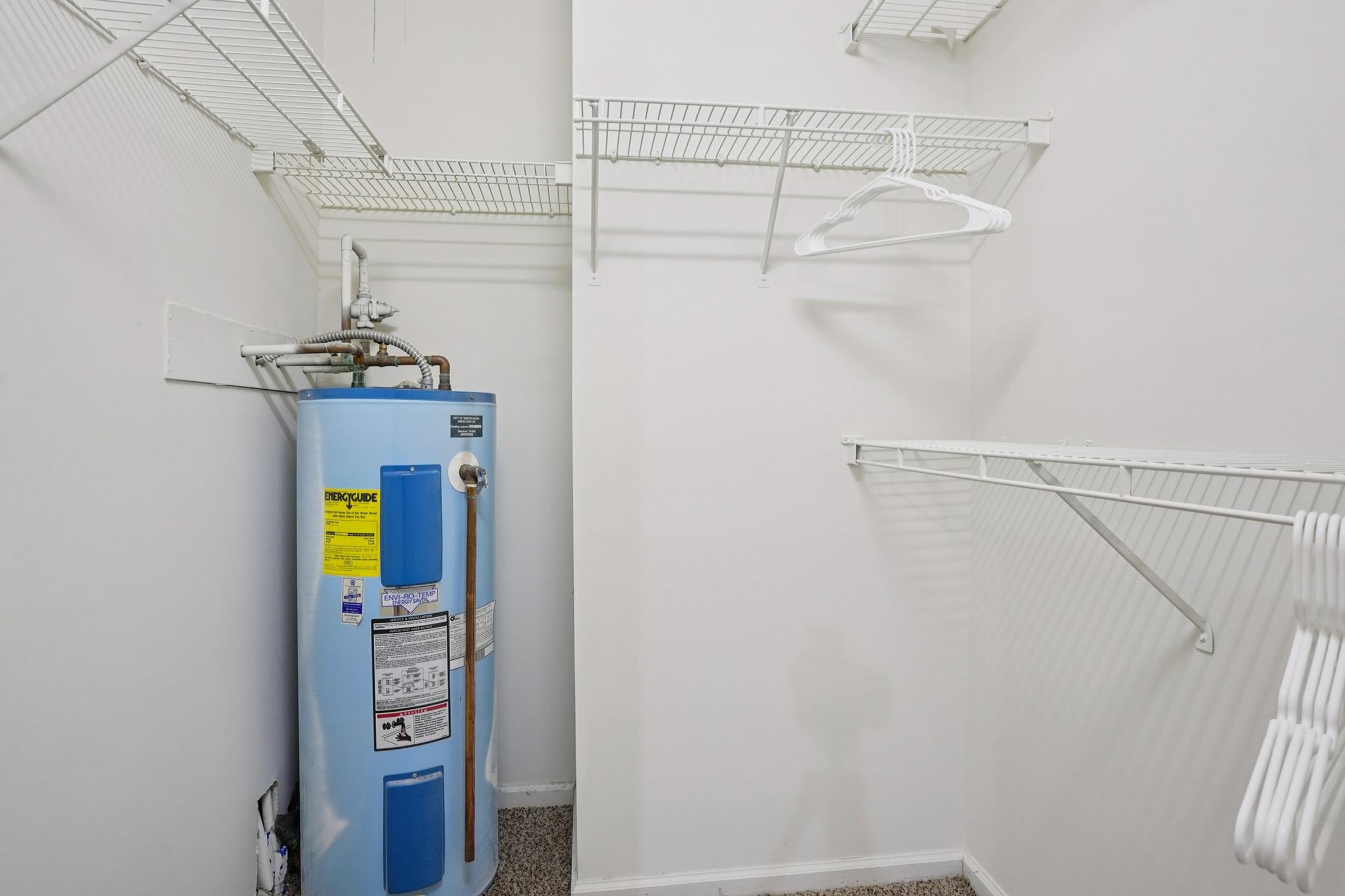 A small closet featuring a blue water heater, several white wire shelves, and a few empty hangers. The walls are painted white, and the floor is carpeted. The shelves provide storage space, but there are no items displayed in the closet.