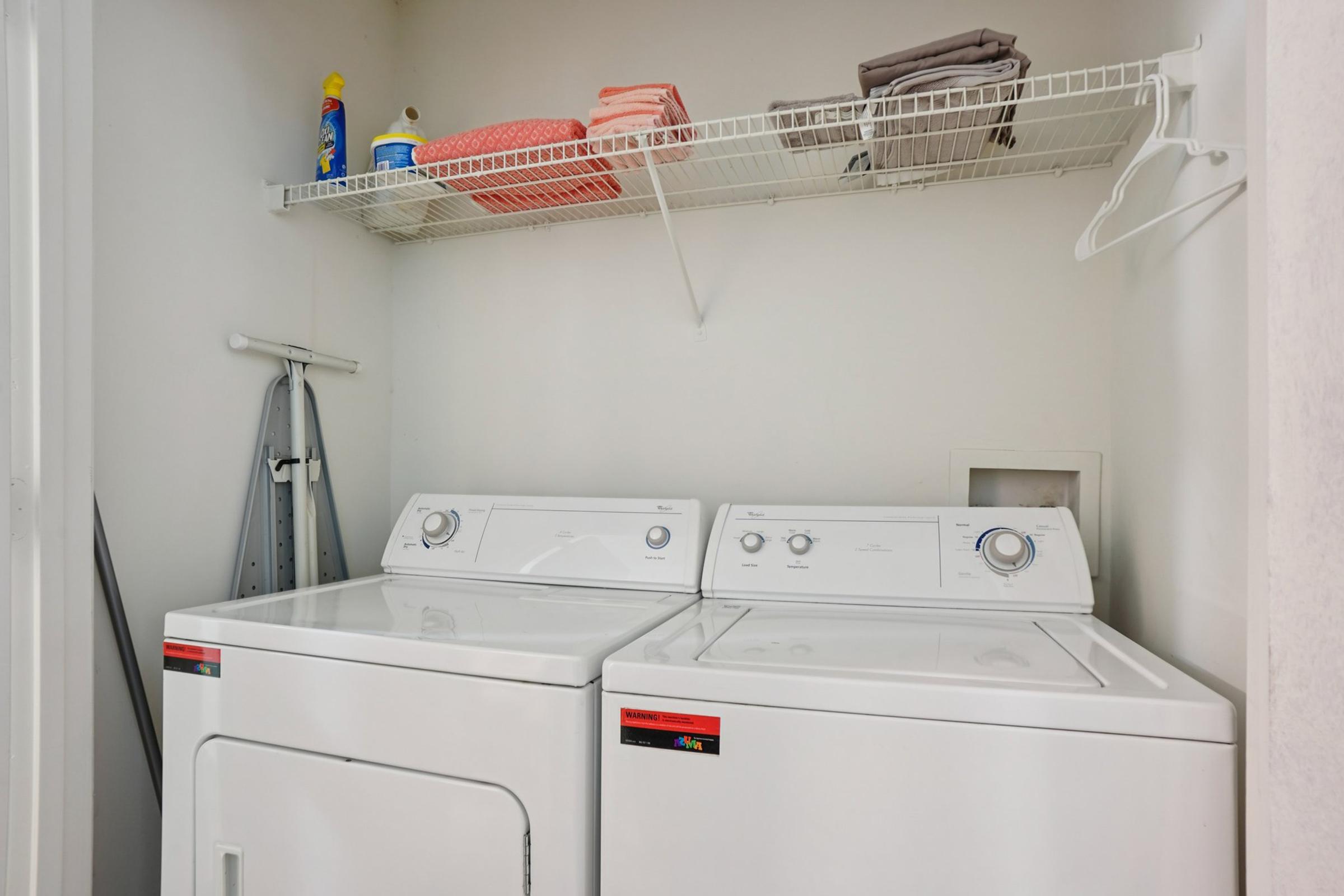 A small laundry room featuring a white washing machine and dryer side by side. Above them is a wire shelf with various laundry supplies, including detergent and stacks of towels. A folding drying rack is leaning against the wall, and the overall space is well-lit and organized.