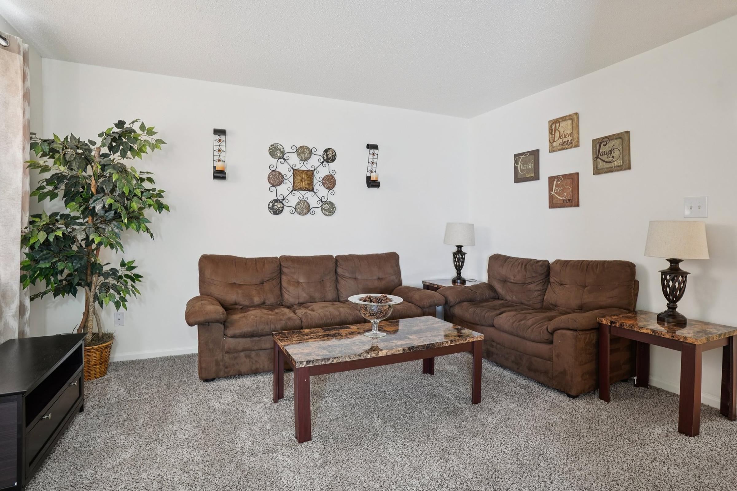 A cozy living room featuring two brown sofas, a wooden coffee table with a decorative bowl, and two side tables with lamps. The walls are adorned with framed art pieces, and there is a potted plant in one corner. The floor is covered with light-colored carpet.