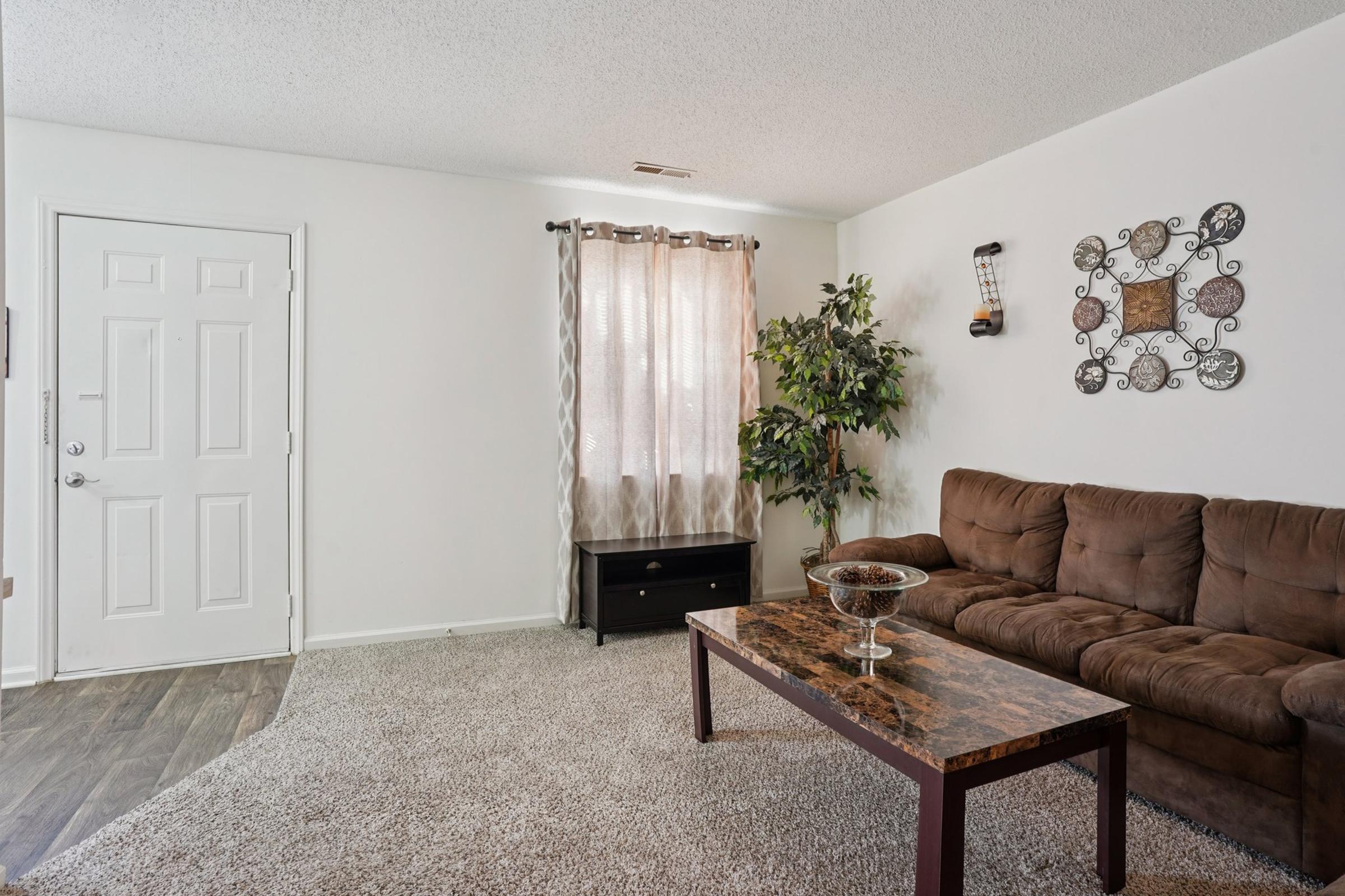 A cozy living room featuring a brown sectional couch, a dark coffee table, and a light-colored rug. There's a decorative wall hanging, a potted plant, and sheer curtains allowing natural light through a window. The room has white walls and a front door visible to the left.