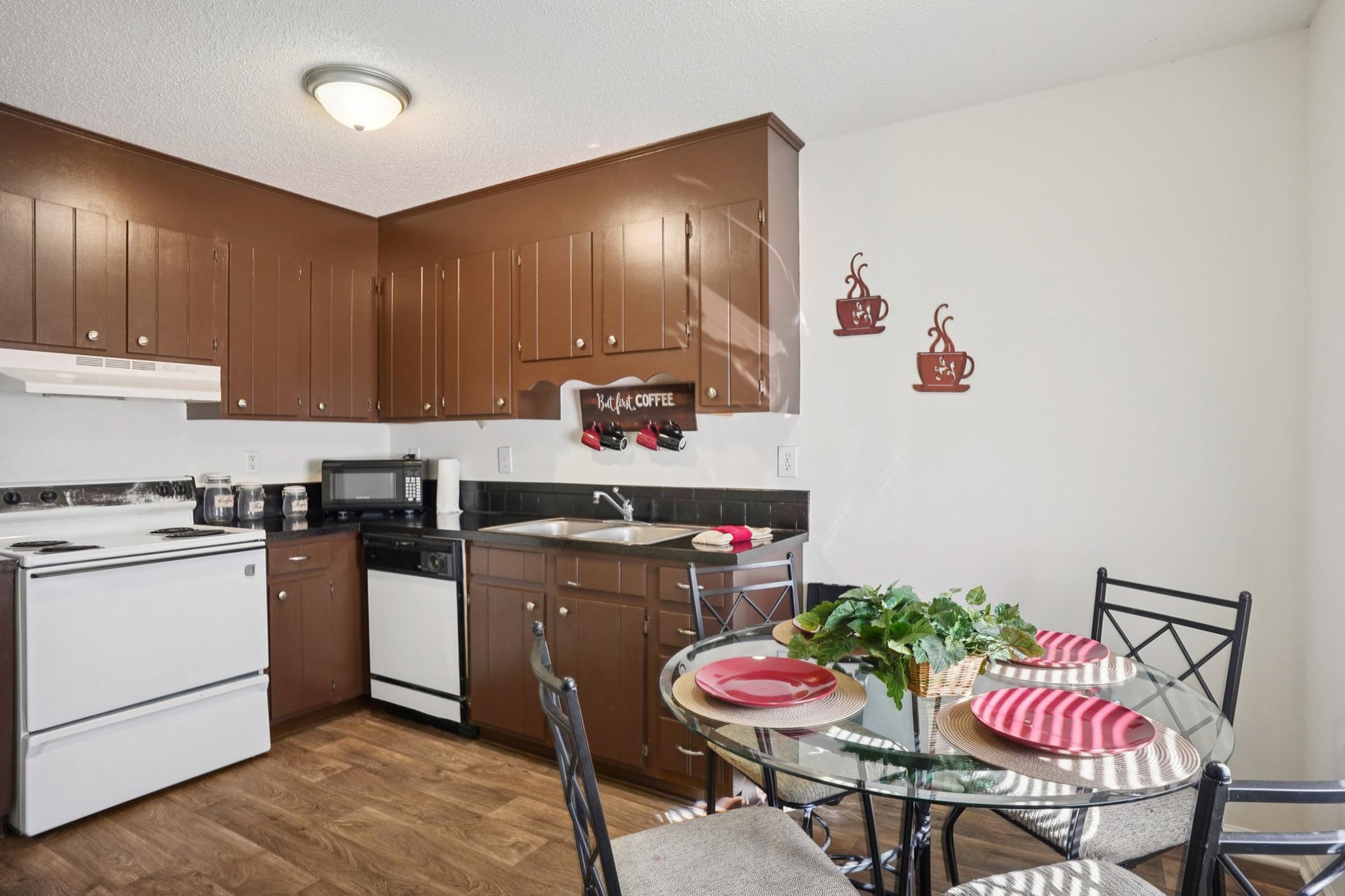 A cozy kitchen featuring dark brown cabinetry, a white stove, and a black microwave. A round glass dining table with black metal chairs is set for two, adorned with red and white plates. Decorative coffee-themed artwork hangs on the wall, adding a warm and inviting atmosphere.