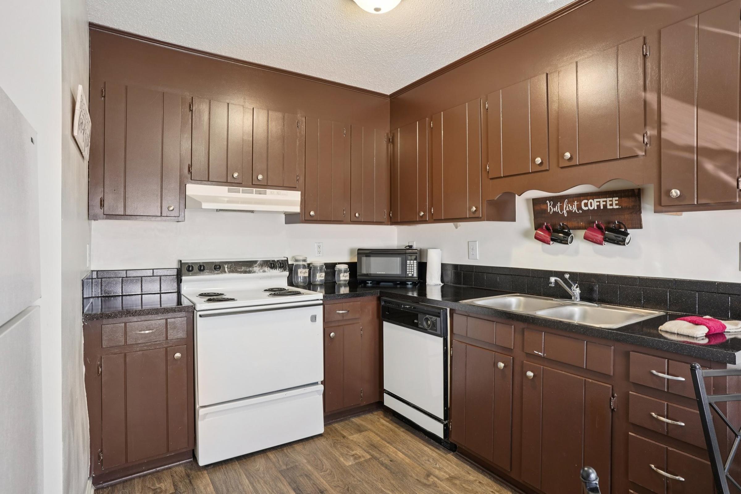Kitchen featuring dark brown cabinets, a white stove, a black refrigerator, and a black sink. The countertop has a coffee-themed decor sign and various kitchen utensils. The flooring is wood-like, and the overall design is modern and inviting.