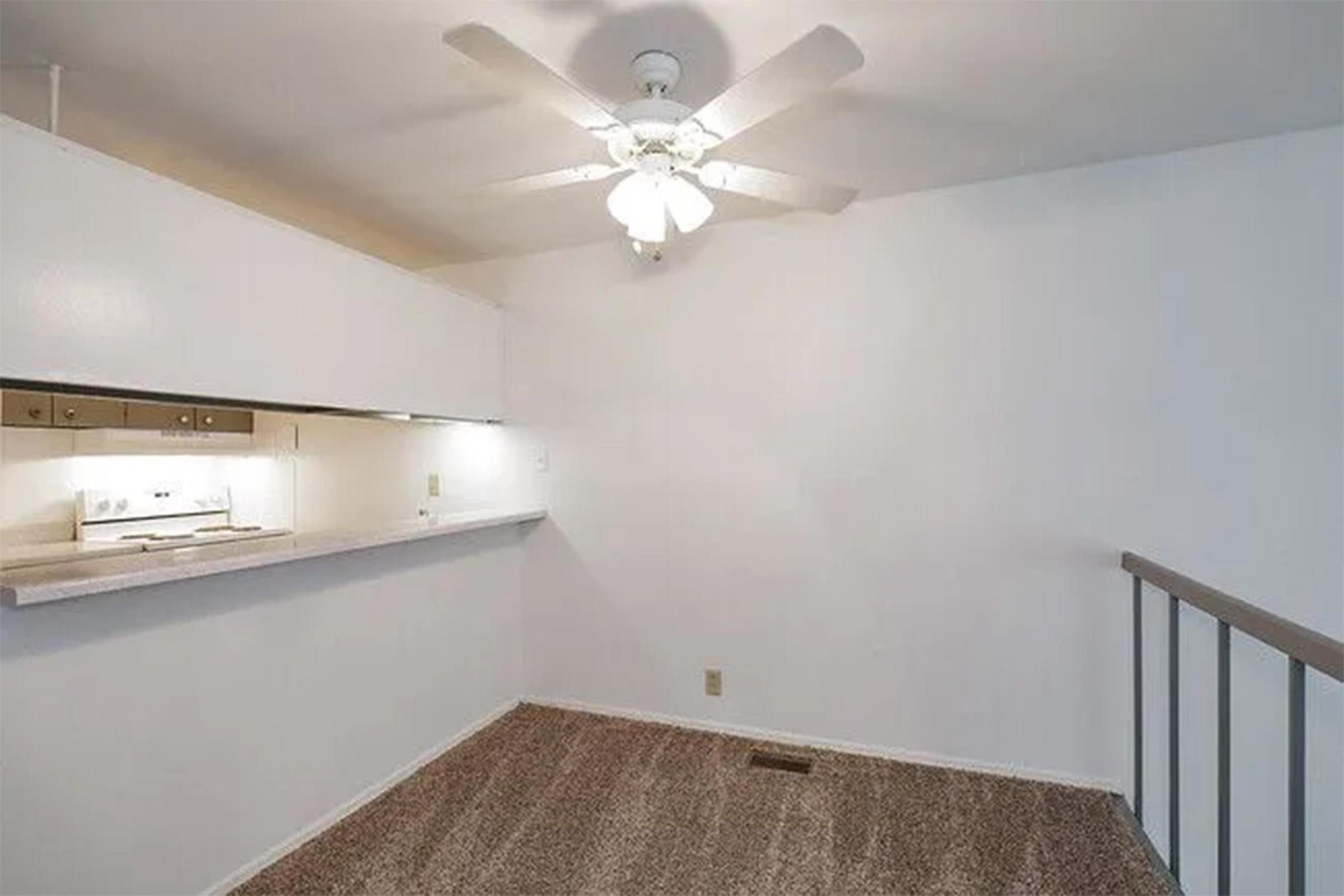 A minimalistic interior space featuring light-colored walls and a ceiling fan. The room includes a staircase railing, a bar-style counter with kitchen fixtures in the background, and beige carpet flooring. Natural light illuminates the area, creating a bright and airy atmosphere.