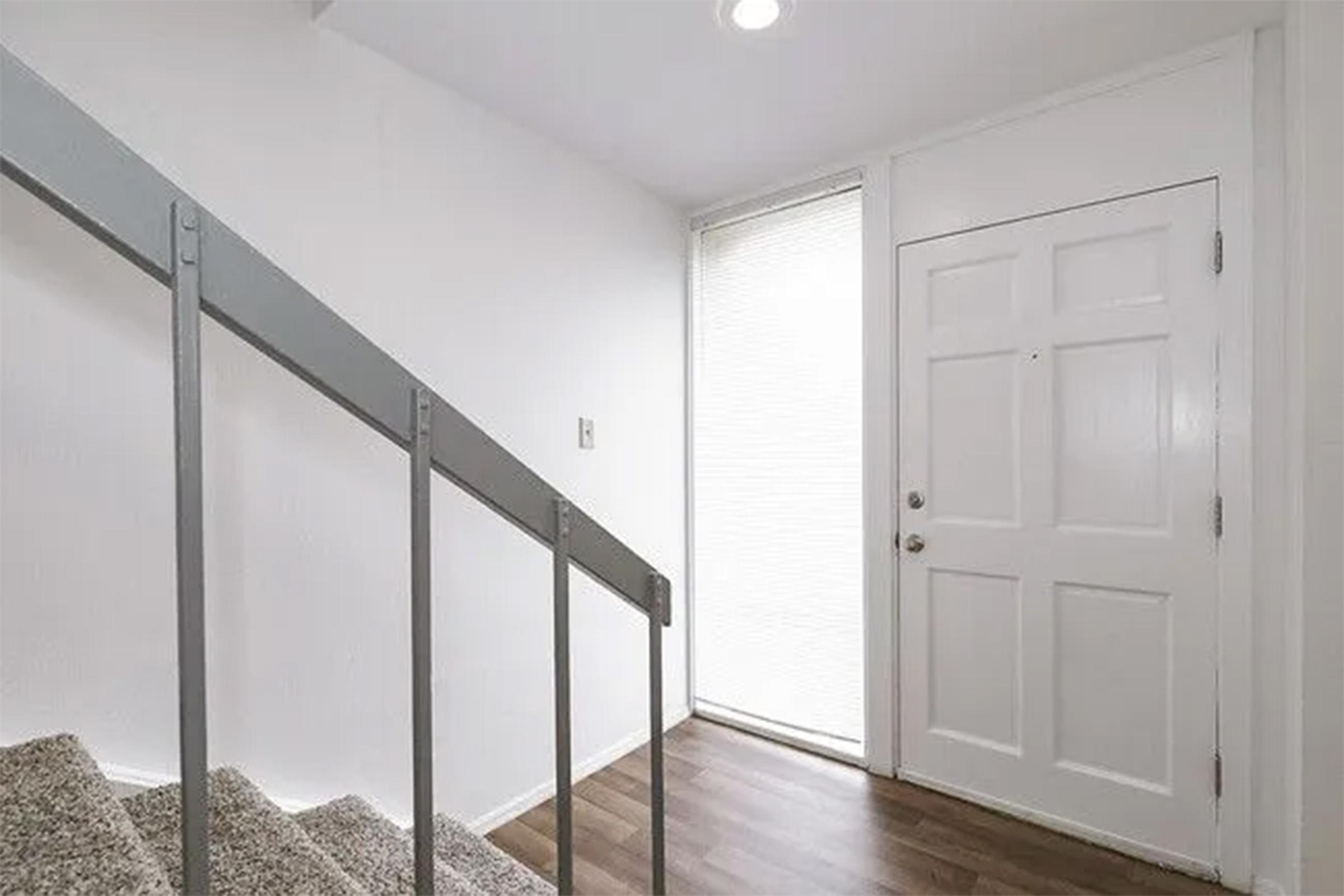 Interior view of a modern entryway featuring a staircase with a metal railing, light-colored walls, and a large front door. Natural light enters through a window beside the door, illuminating the space. The flooring is a wooden surface, creating a clean and inviting atmosphere.