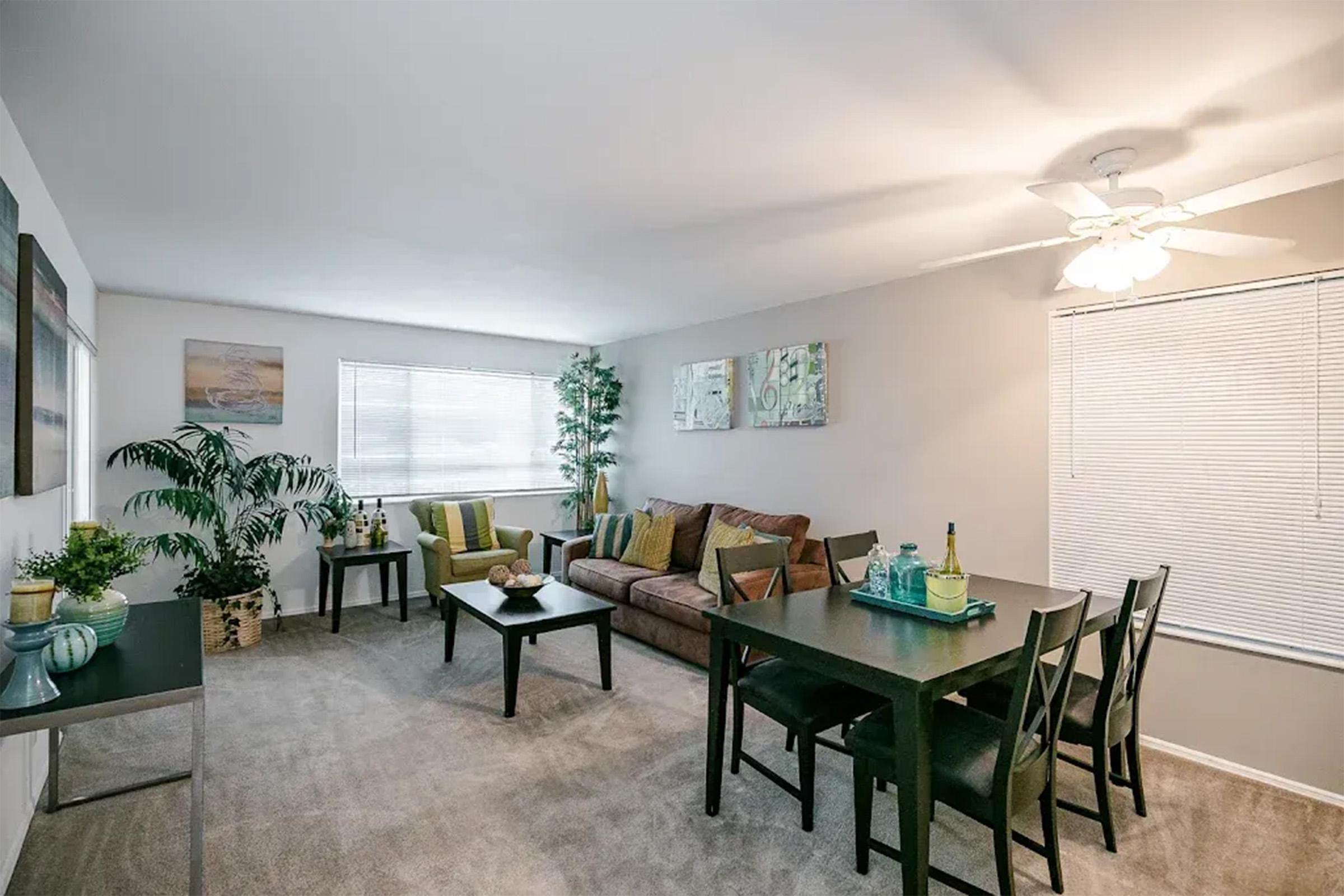 A cozy living and dining area featuring a brown sofa, a dark dining table with chairs, and decorative plants. The walls are adorned with artwork, and natural light streams through the windows, creating a warm atmosphere. The room is carpeted and well-lit with a ceiling fan.