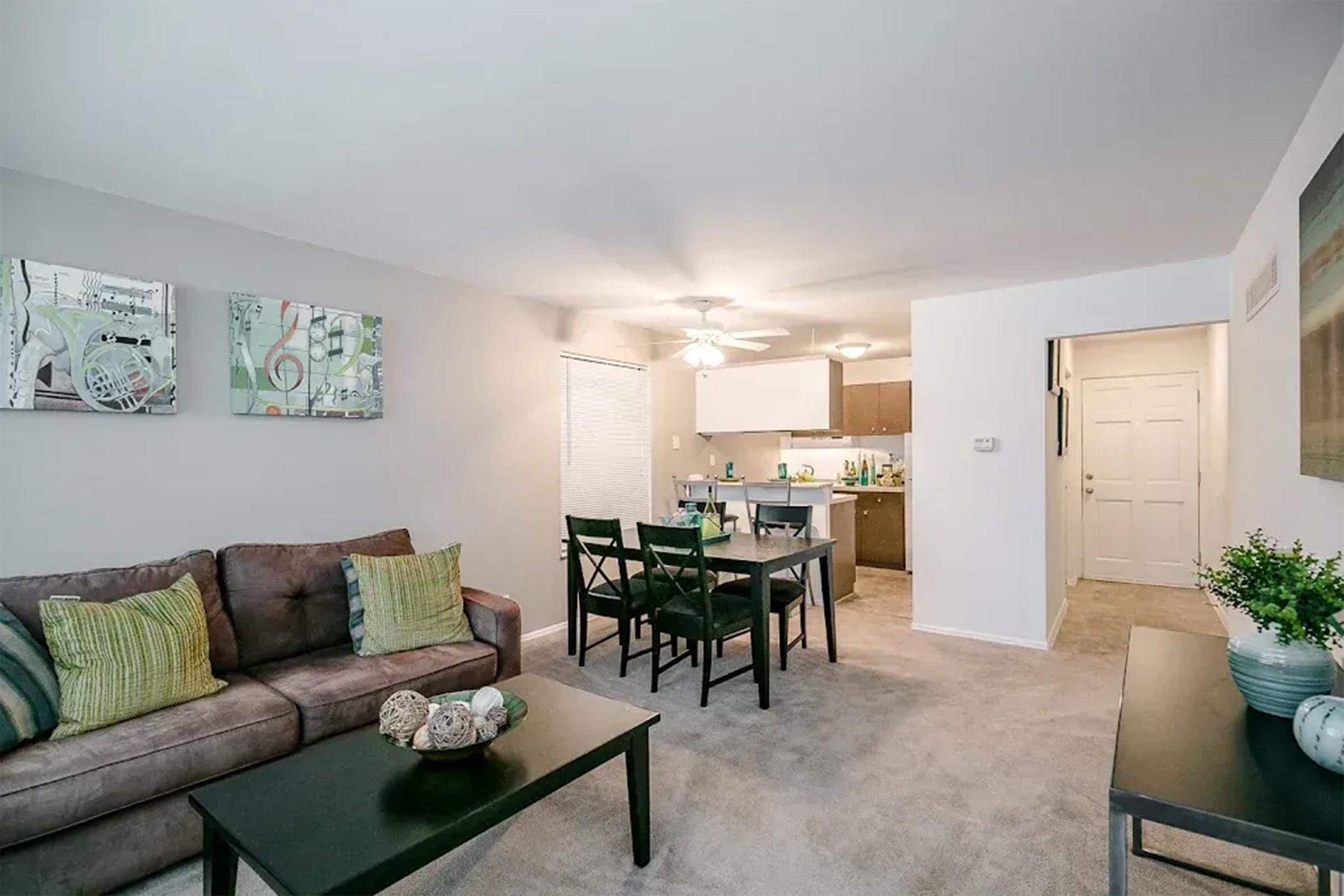 A cozy living room featuring a brown sofa with green accent pillows, a black coffee table, and a dining area with a table and chairs. The walls are adorned with abstract artwork, and the space is well-lit by a ceiling fan. A doorway leads to a kitchen area in the background.